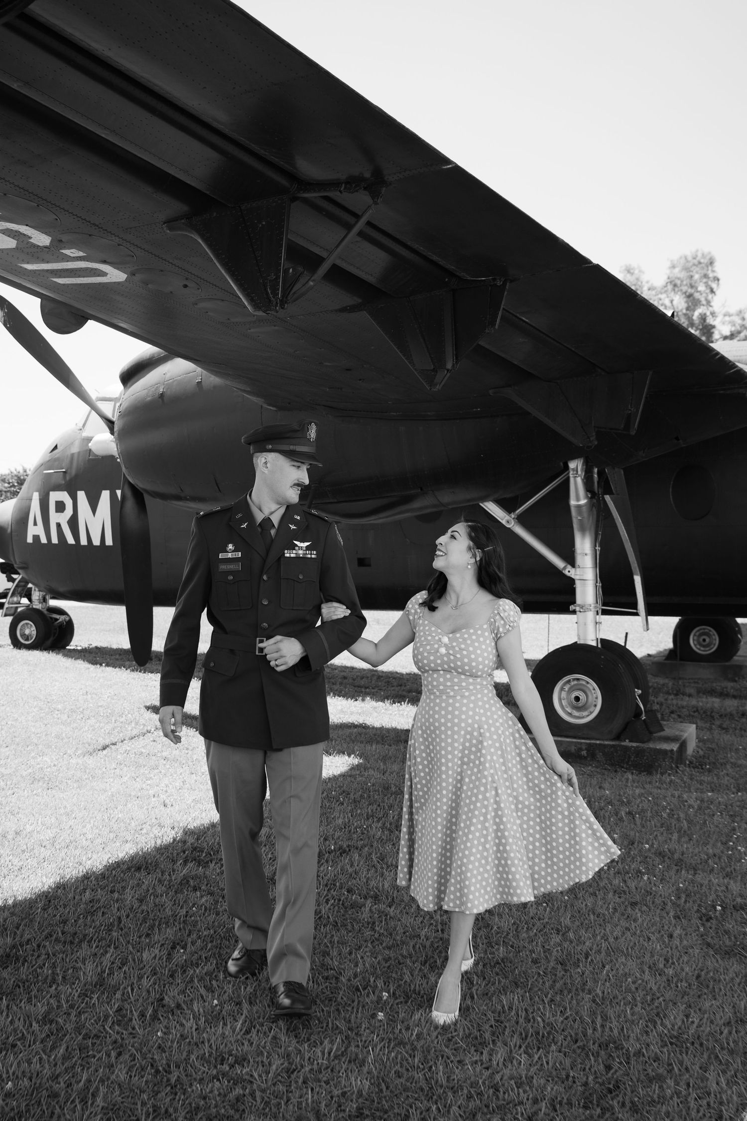 Black and white photo of a soldier in uniform walking with a woman in a polka dot dress beside a vintage Army aircraft.