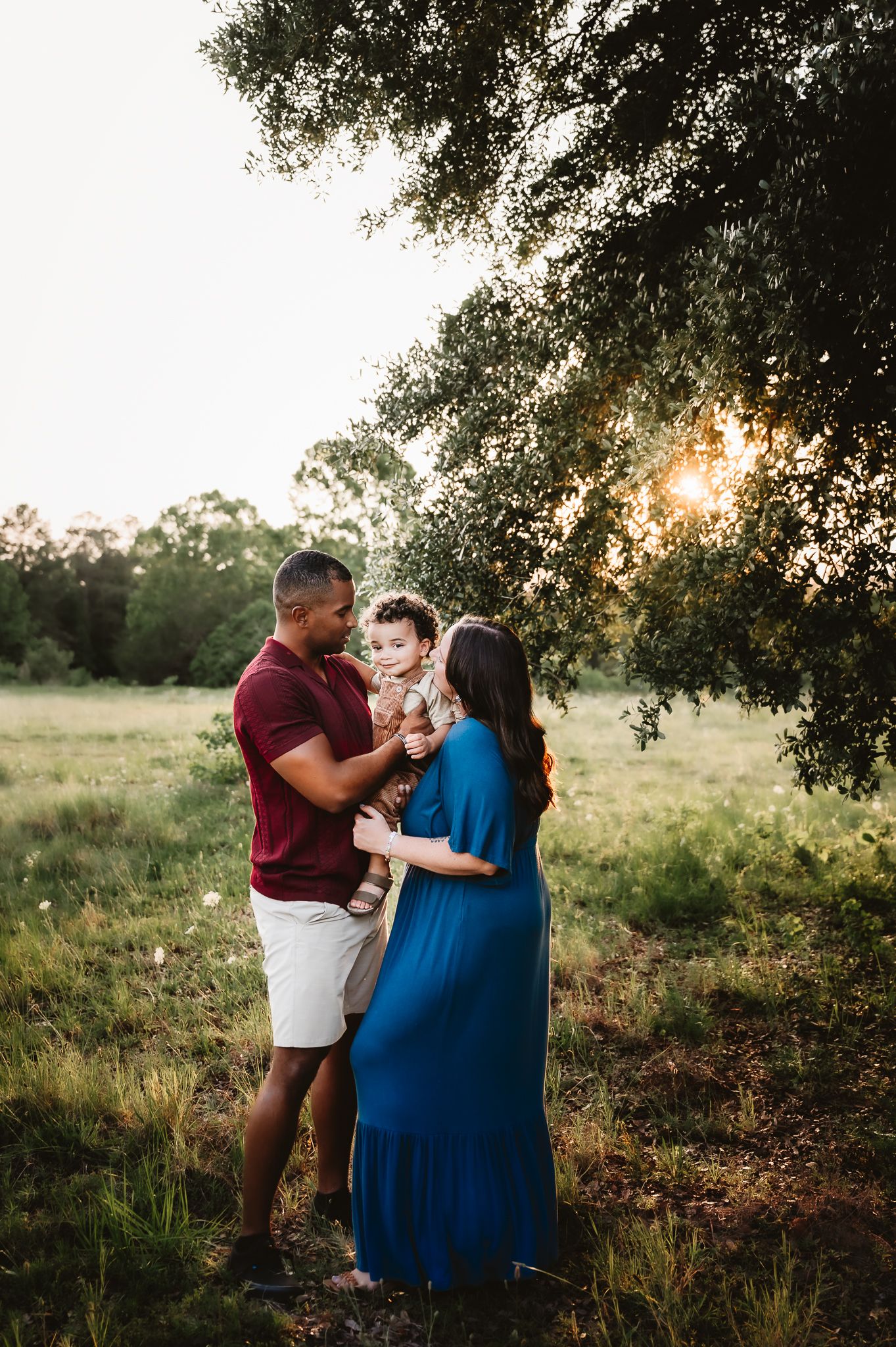 A couple embraces tenderly under tree branches at sunset in a serene countryside setting.