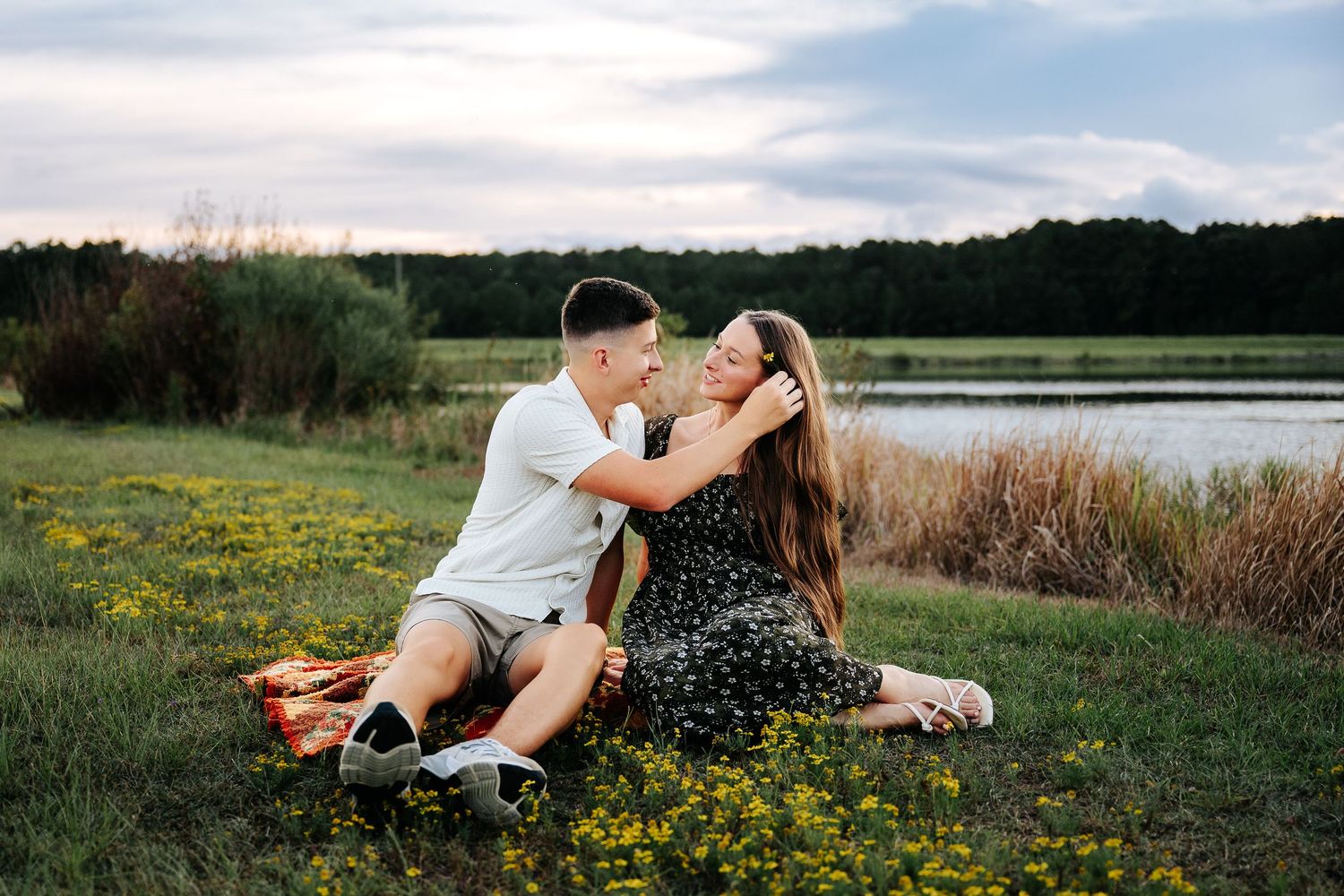 Couple sitting in a field of wildflowers by a lake at sunset, sharing a romantic embrace.