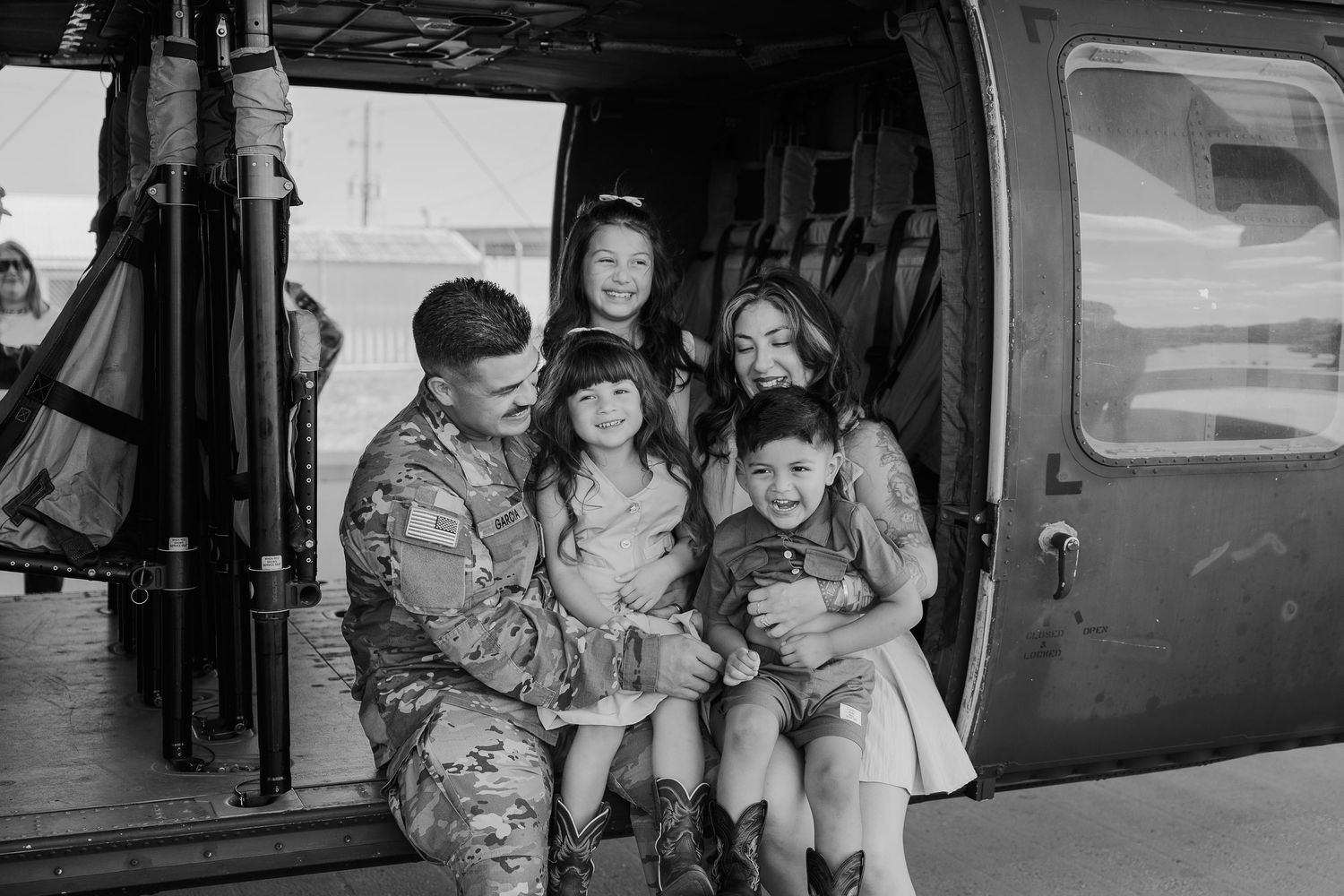 Military soldier in uniform sits with his family of four children in the open door of a helicopter.