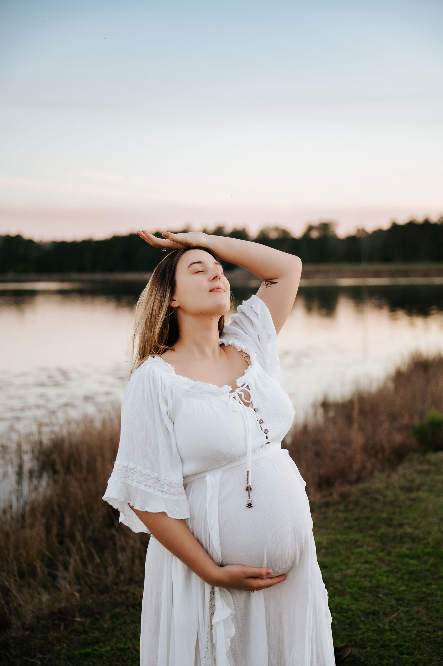 A pregnant woman in a flowing white dress poses gracefully by a serene lake at sunset.