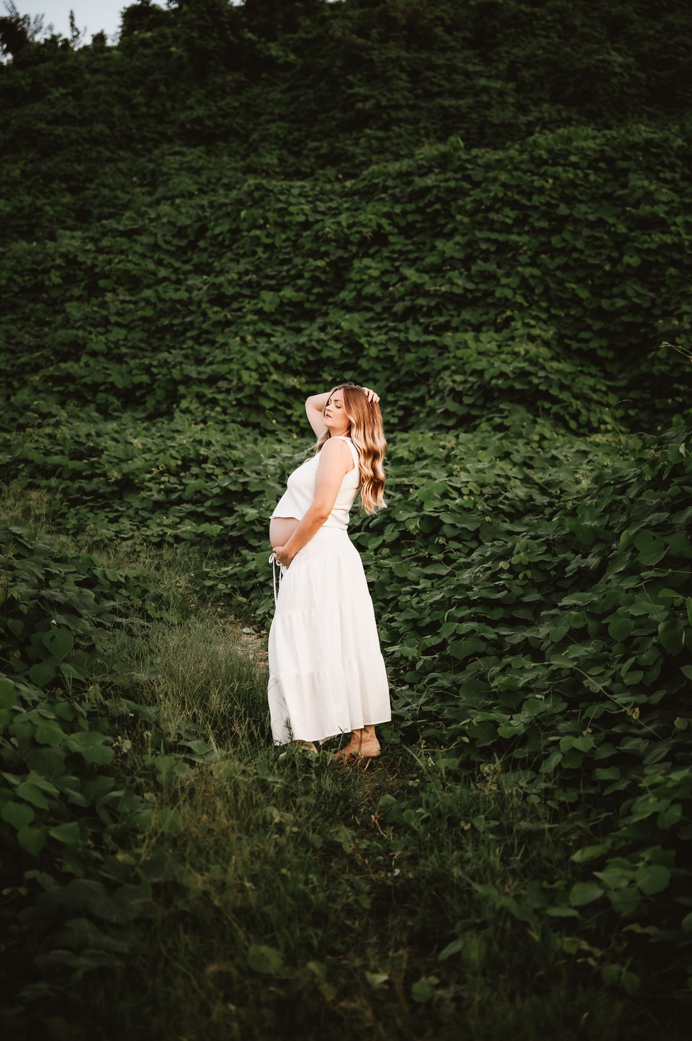 A person wearing a flowy white dress stands contemplatively at the edge of a lush green forest pathway.