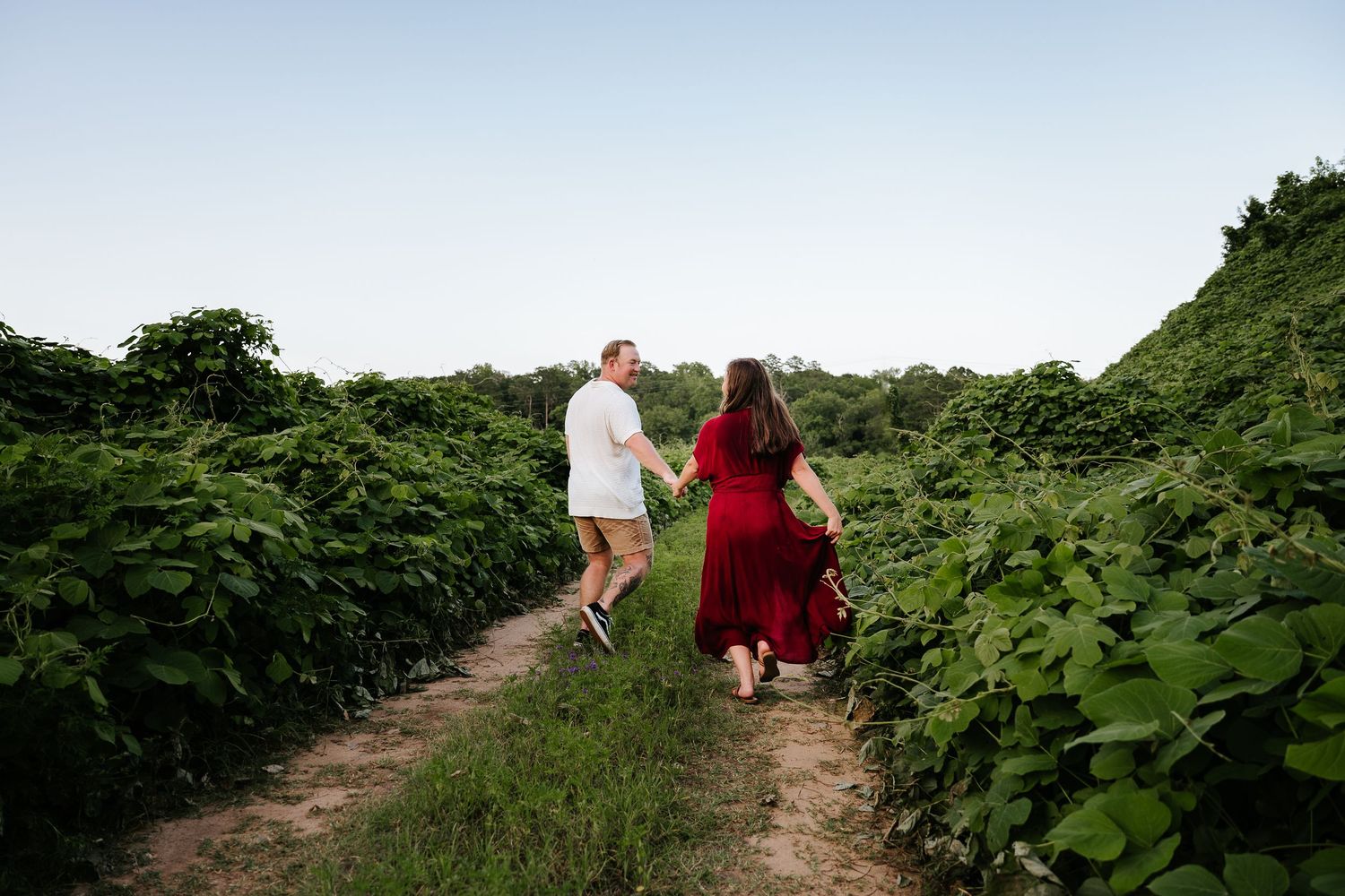 Couple holding hands walking through lush green garden path in golden hour light, woman in red dress.