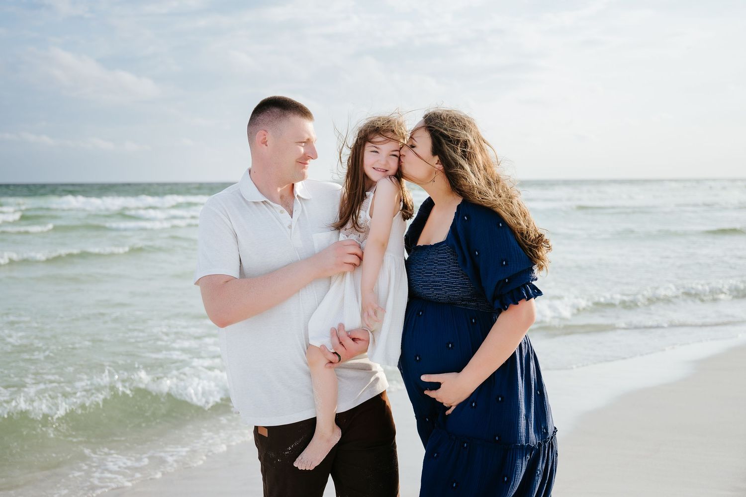 Pregnant woman in navy dress with husband and daughter on sandy beach with turquoise ocean waves in background.