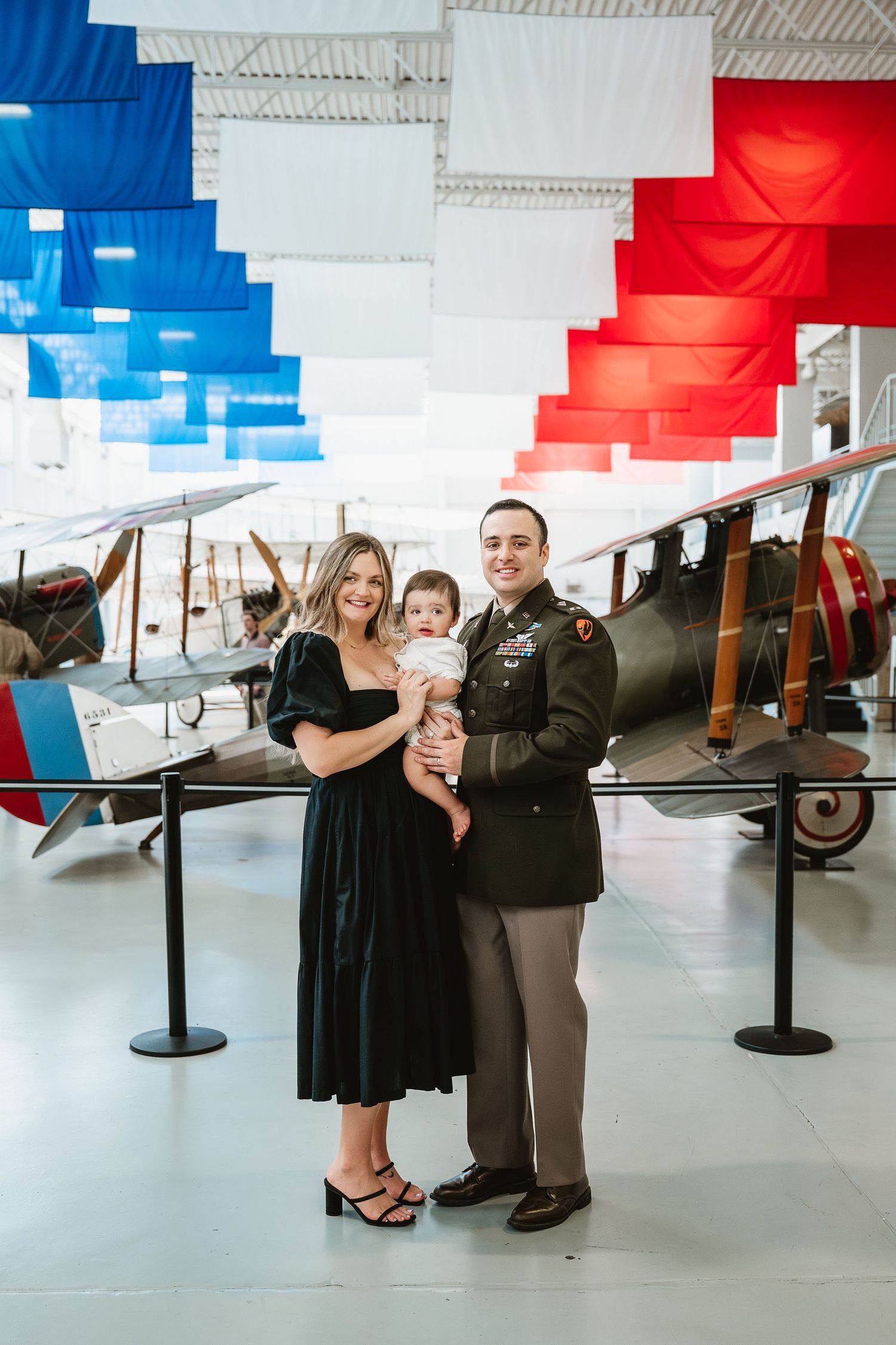 Military family poses with baby in aviation museum, red white and blue banners and vintage aircraft displayed behind them.