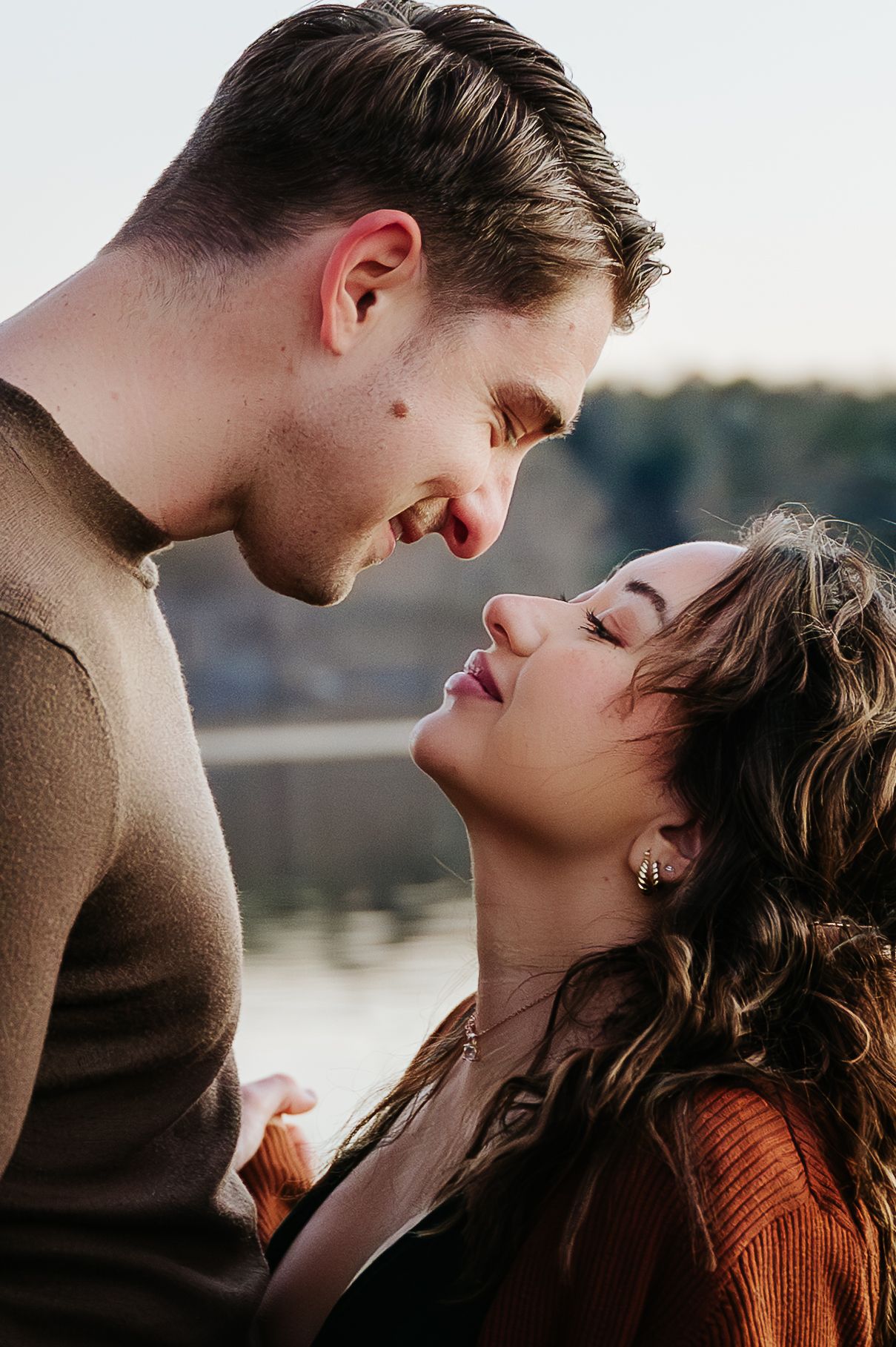 Romantic couple in warm sweaters share an intimate moment by the water during golden hour.