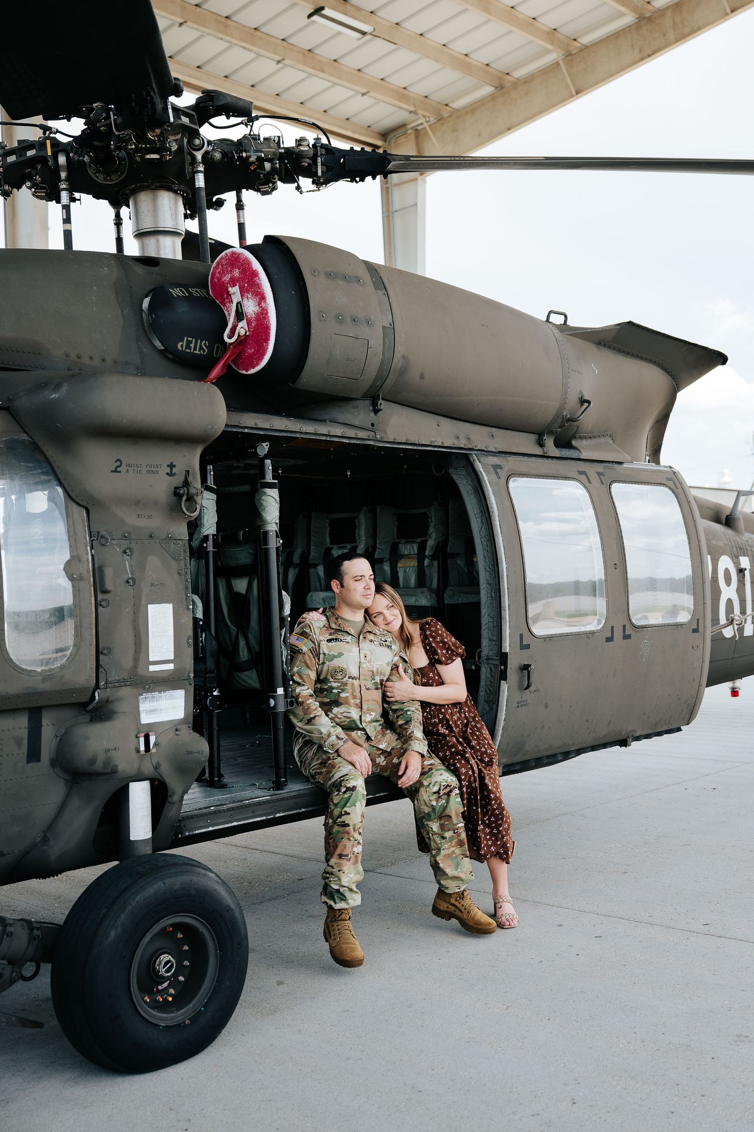 A soldier in camouflage uniform embraces a woman in a floral dress beside a military helicopter in a hangar.