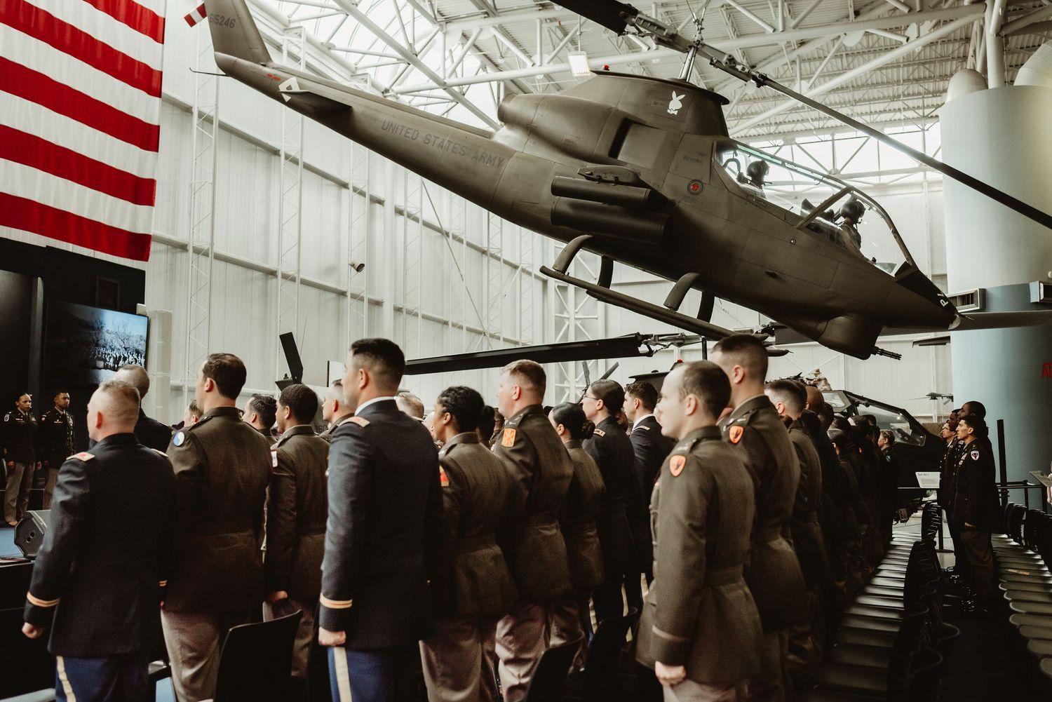 Military personnel gather near an Apache helicopter with American flag in background.