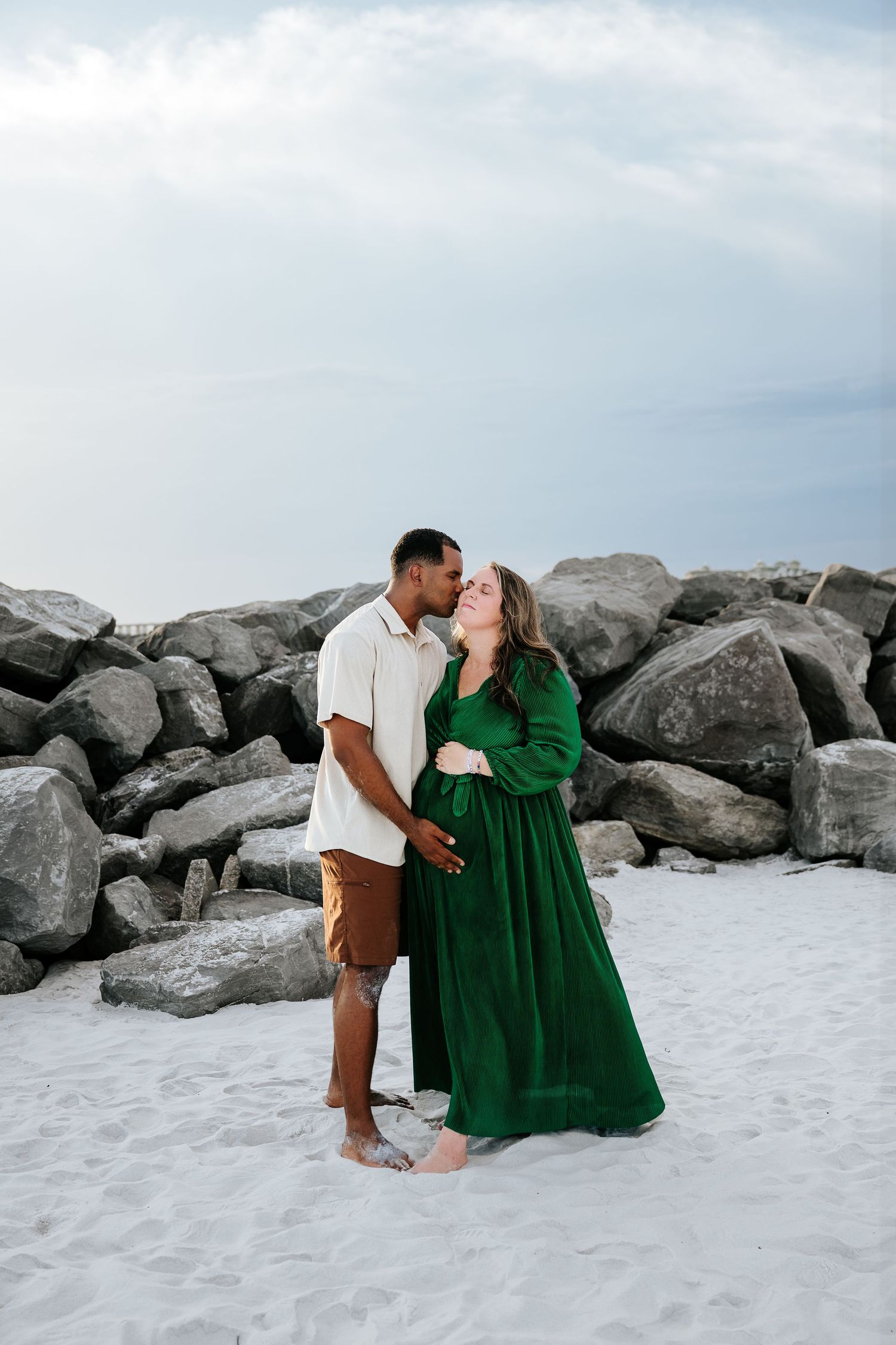 Pregnant couple sharing a kiss on a white sand beach with rocky backdrop, woman in elegant green maternity dress.