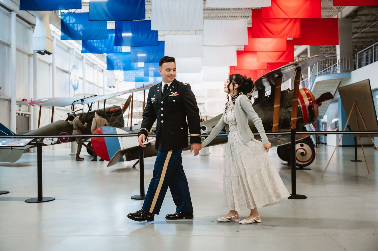 A couple walks hand in hand through an aviation museum with vintage aircraft displays and red and blue banners overhead.