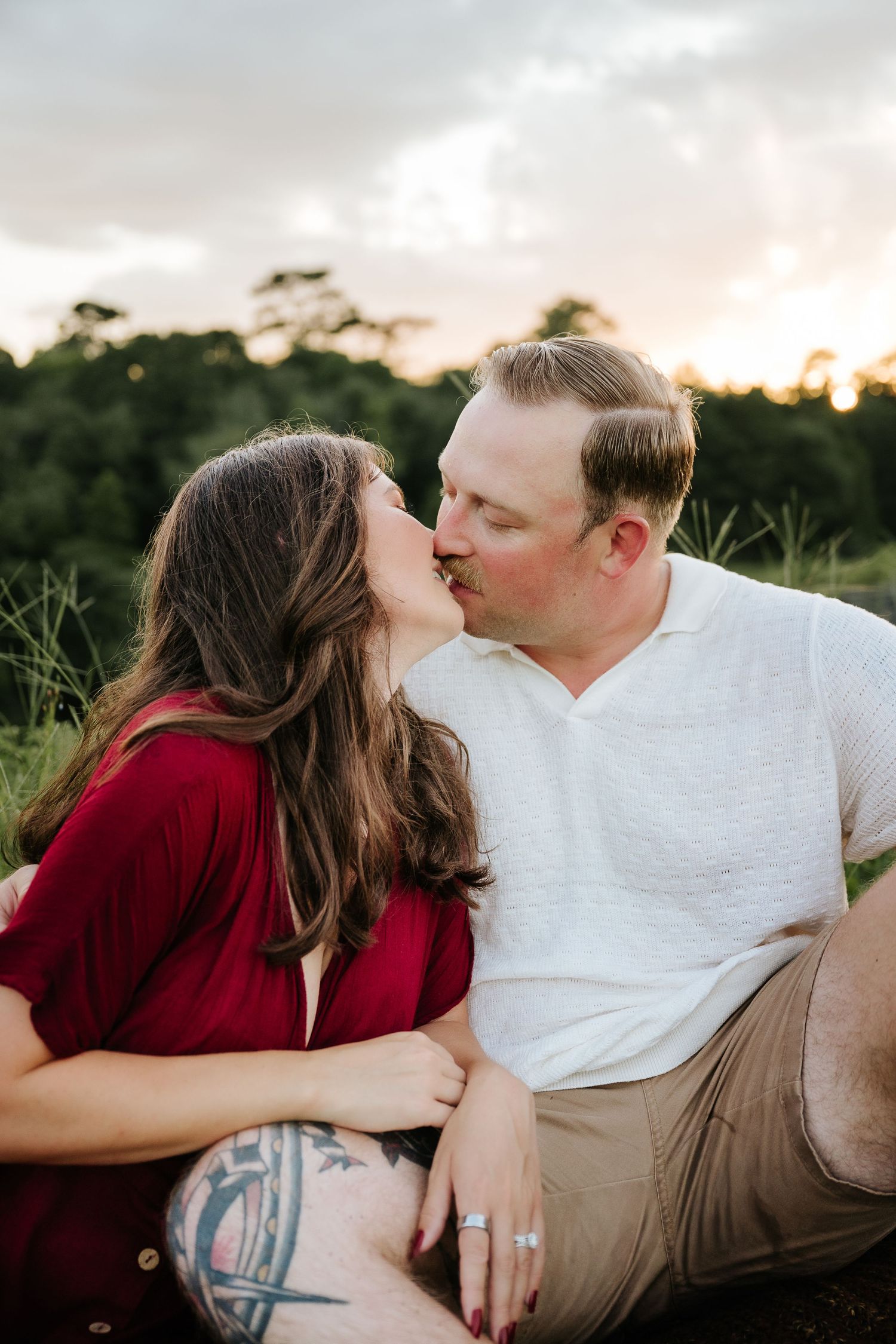 Couple sharing a romantic kiss at sunset in a green field, woman in red top with tattoo, man in white shirt.