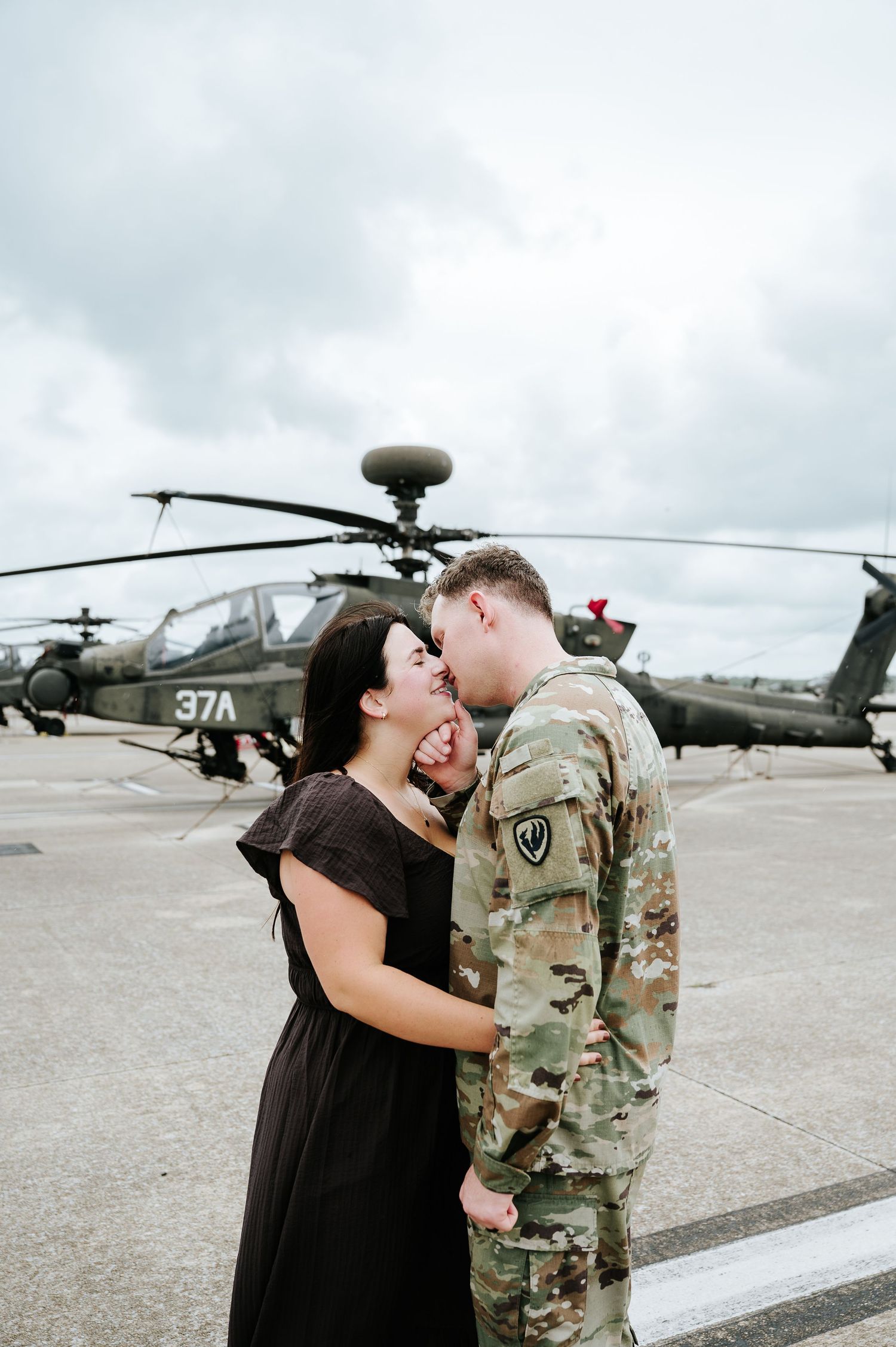 A soldier in camouflage uniform shares a romantic kiss with a woman in a black dress in front of a military helicopter.