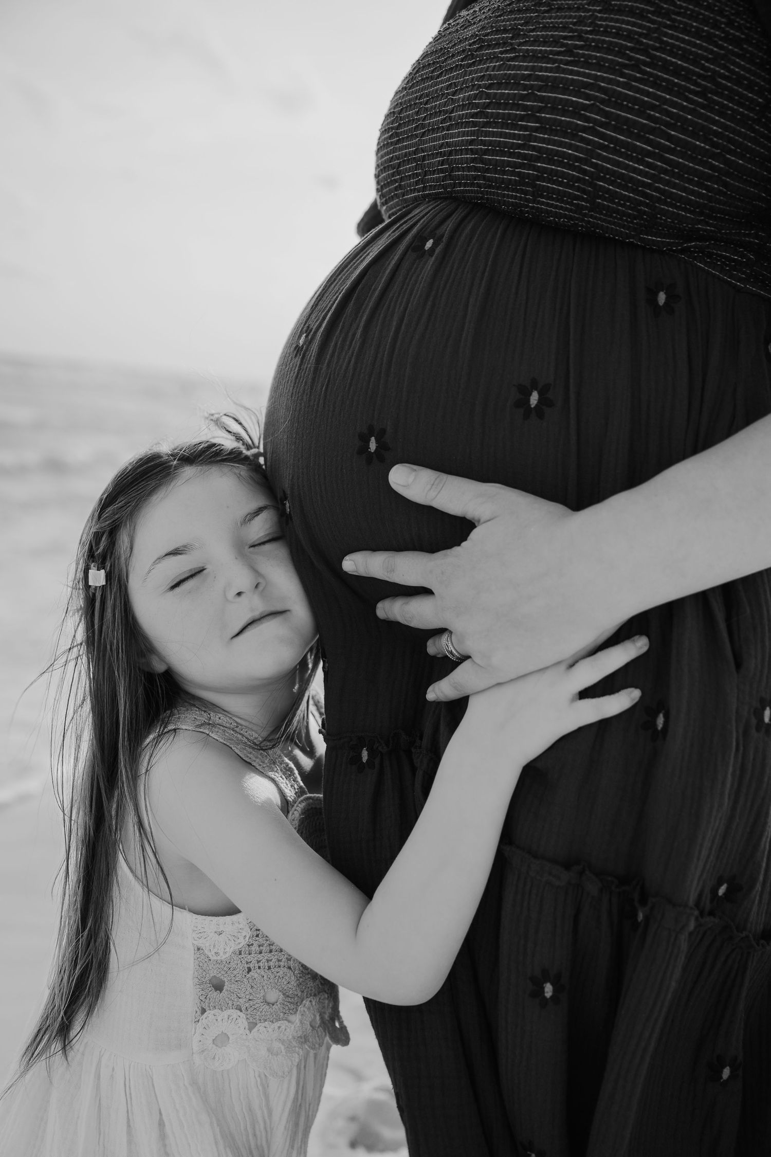 Black and white photo of a young girl hugging her pregnant mother's belly on a beach.