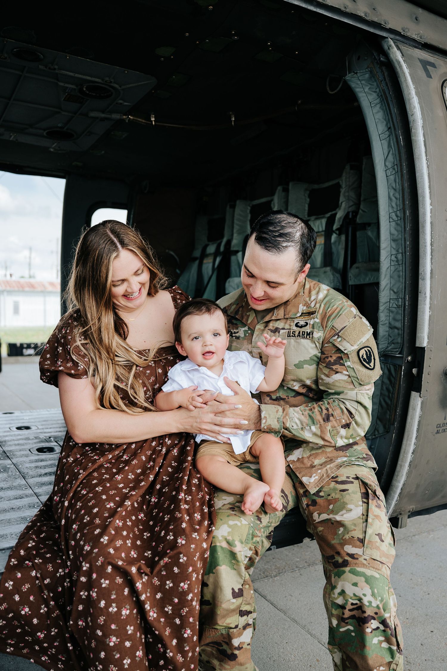 Army soldier in camo uniform sits with wife in brown floral dress and baby by military helicopter.