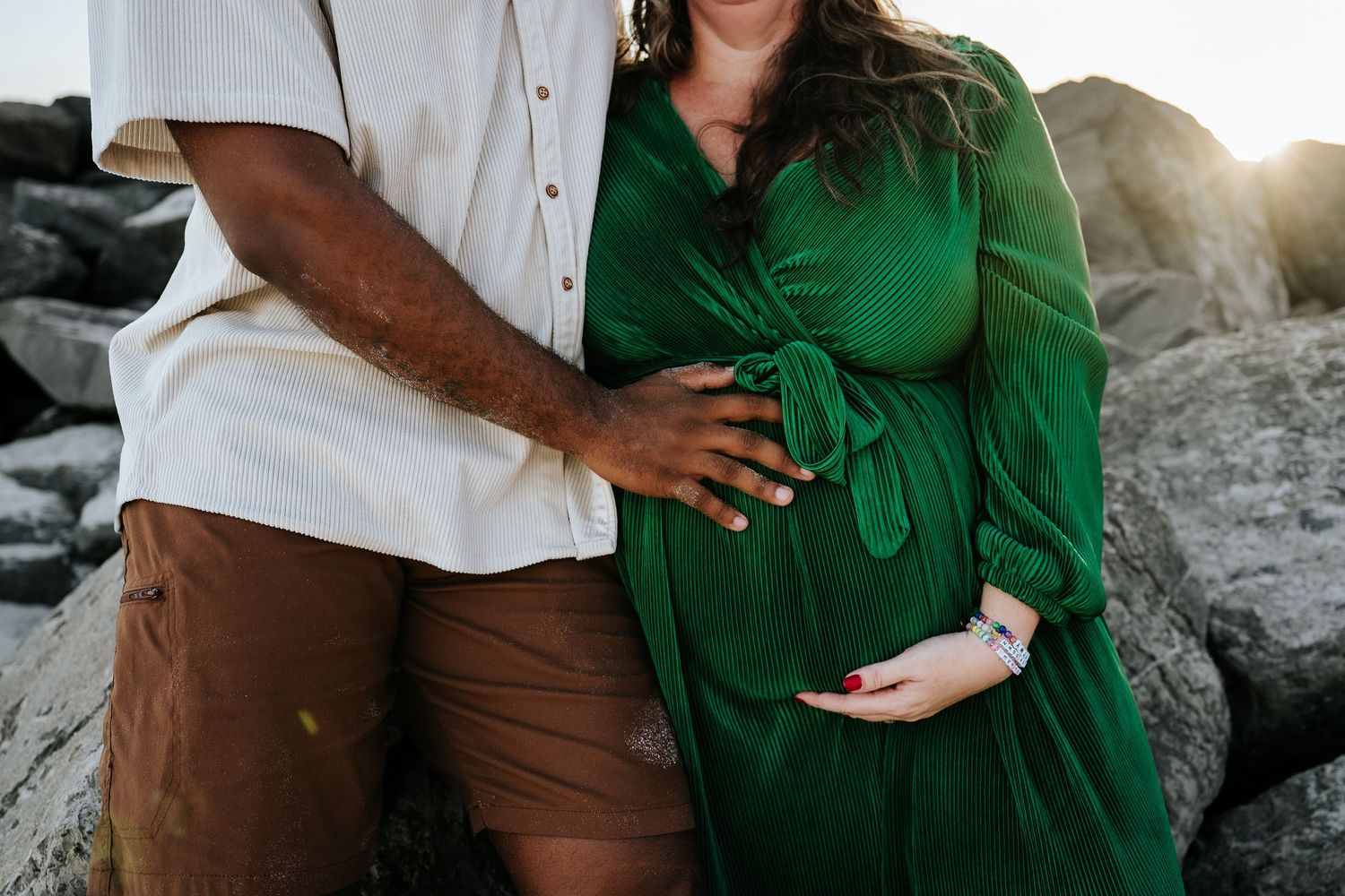 Pregnant woman in green dress with partner touching her baby bump outdoors near rocks during golden hour.