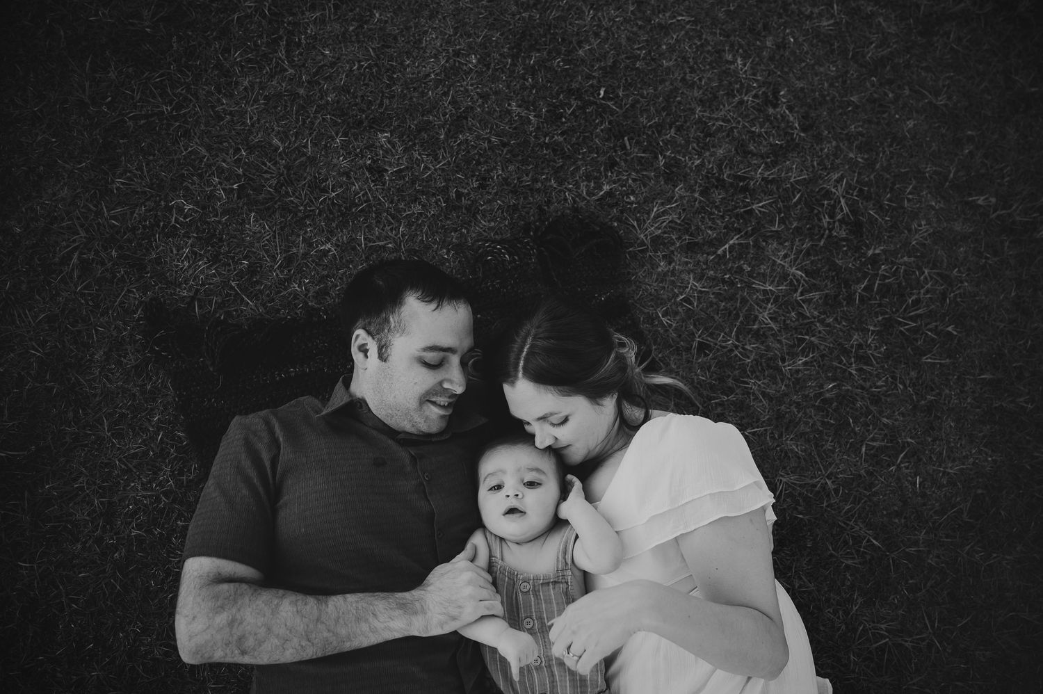 Black and white photo of a couple lying on grass with their baby, sharing a tender family moment outdoors.