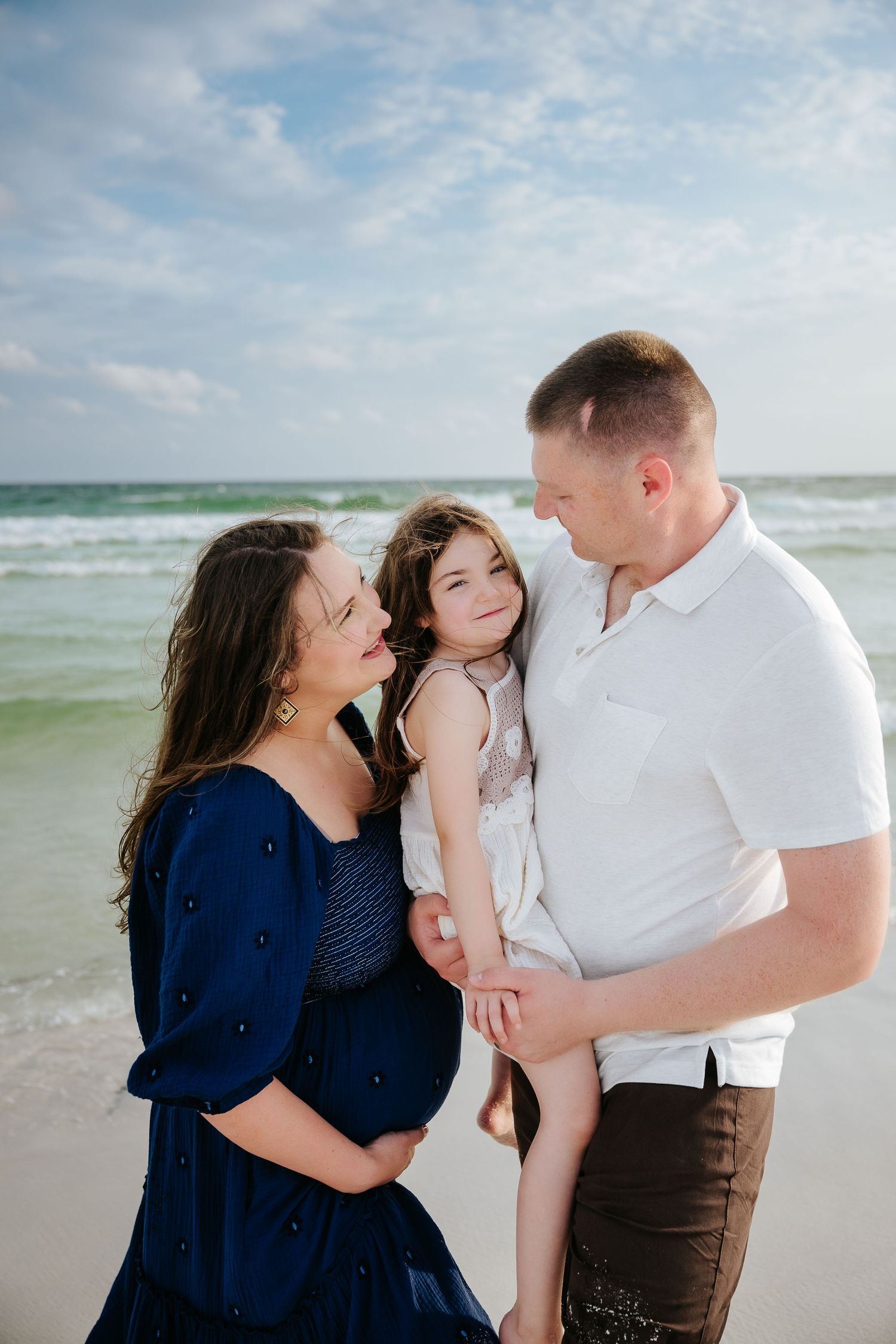 Pregnant woman in blue dress and man in white shirt hold young girl on a sunny beach with turquoise waves.