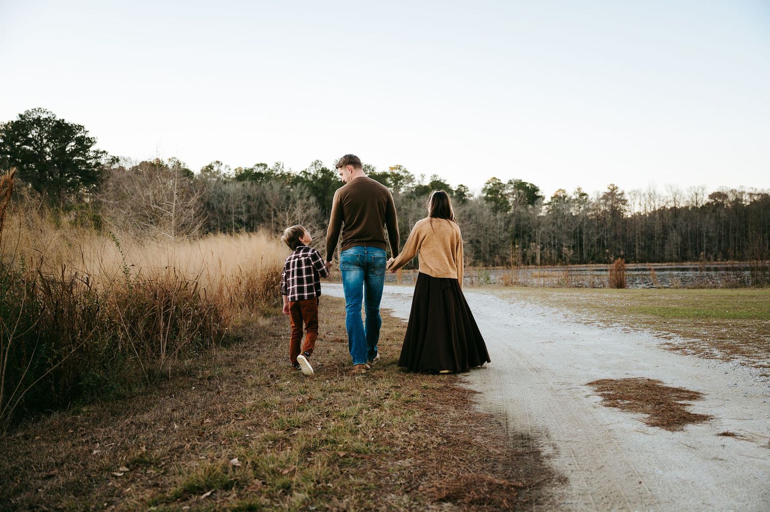 Three people walk together down a dirt path surrounded by tall grass and trees on a winter afternoon.