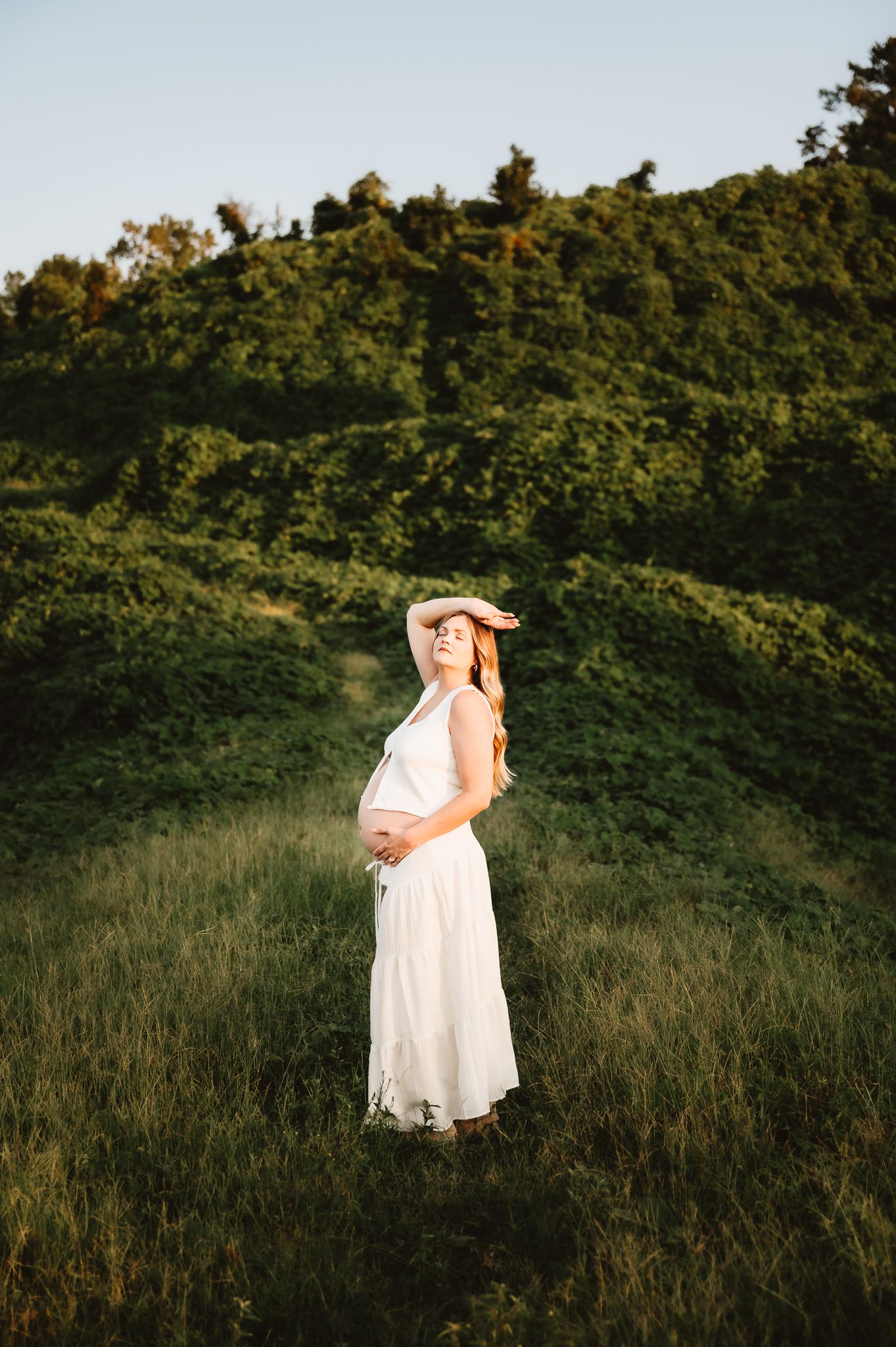 A person in a flowing white dress stands in a golden field at sunset with lush green trees in the background.