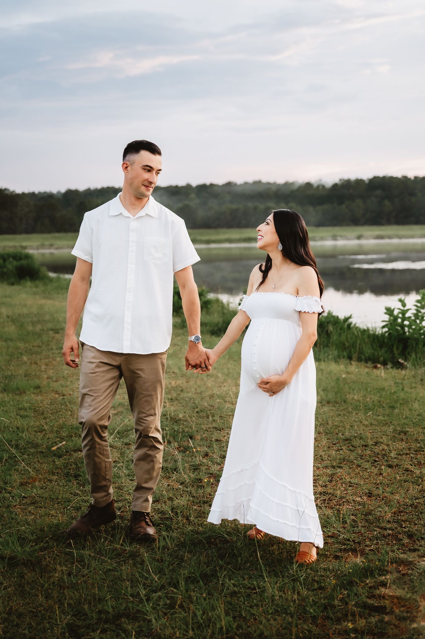 A couple holding hands walks together in a grassy field near a lake during a romantic outdoor photoshoot.
