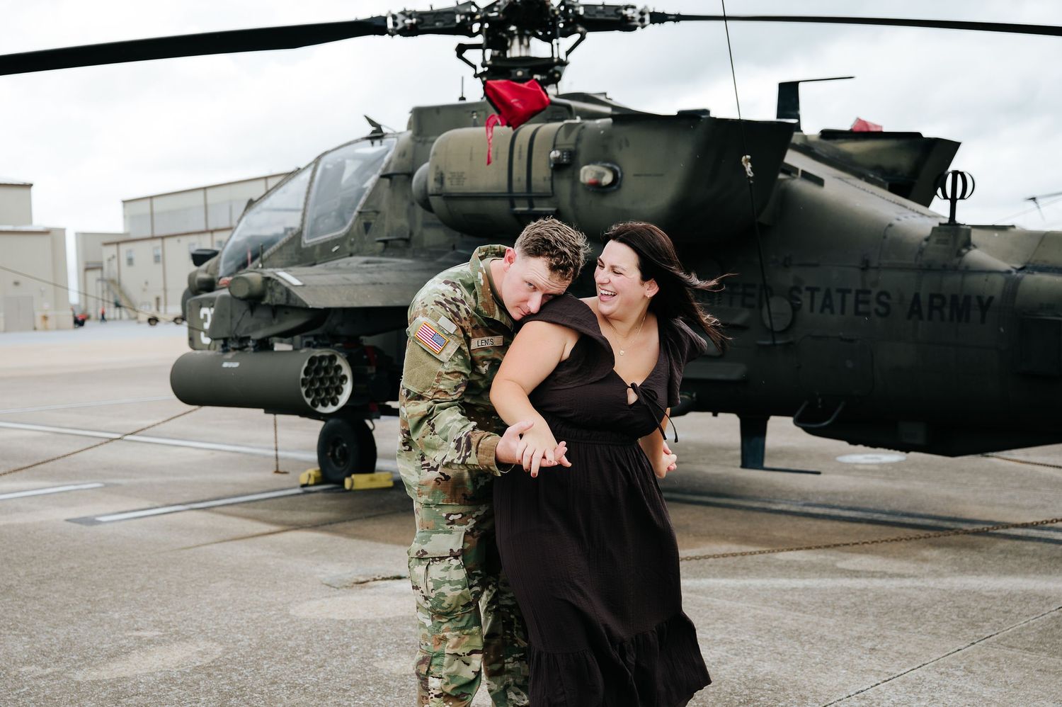 Soldier in camouflage uniform embracing a woman in black dress in front of a U.S. Army helicopter.