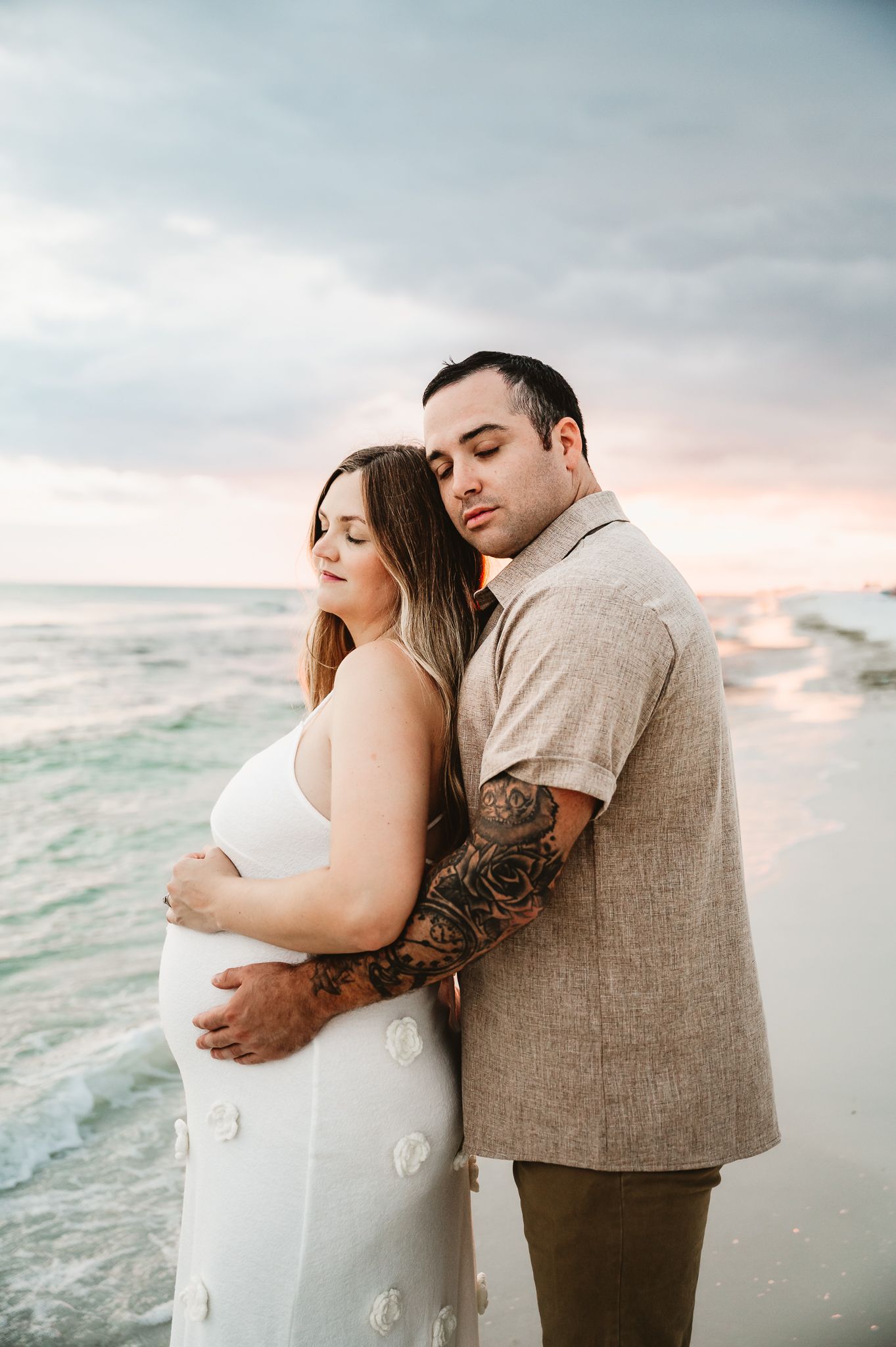 A romantic couple embraces on a cloudy beach at sunset with waves rolling in behind them.