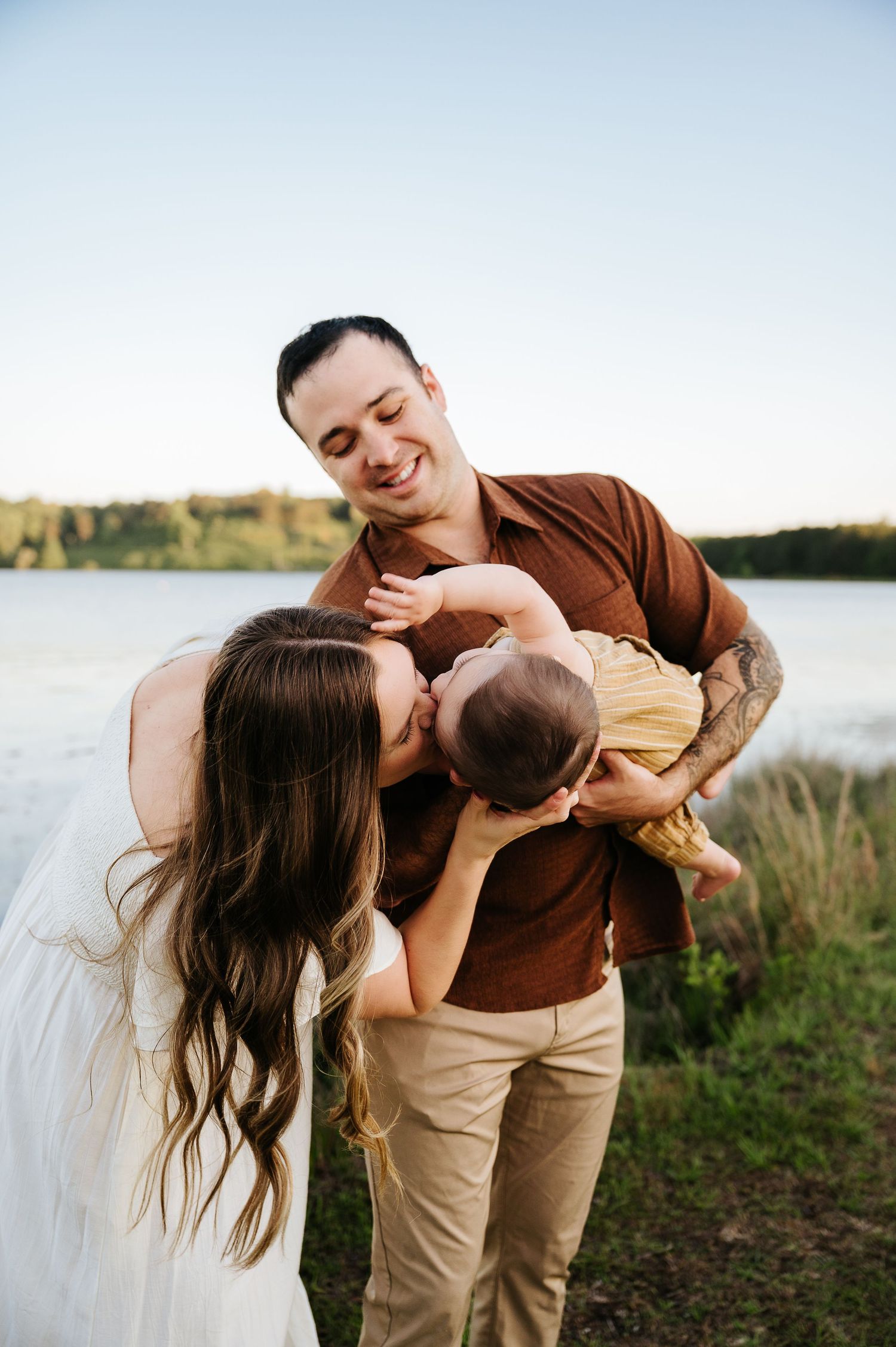 A happy young family outdoors, father holding baby while mother kisses infant near a serene lake at golden hour.