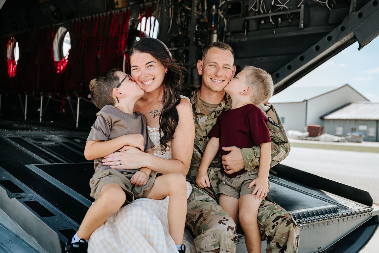 Military family smiling together on aircraft, two young children kissing parents in camouflage uniform.