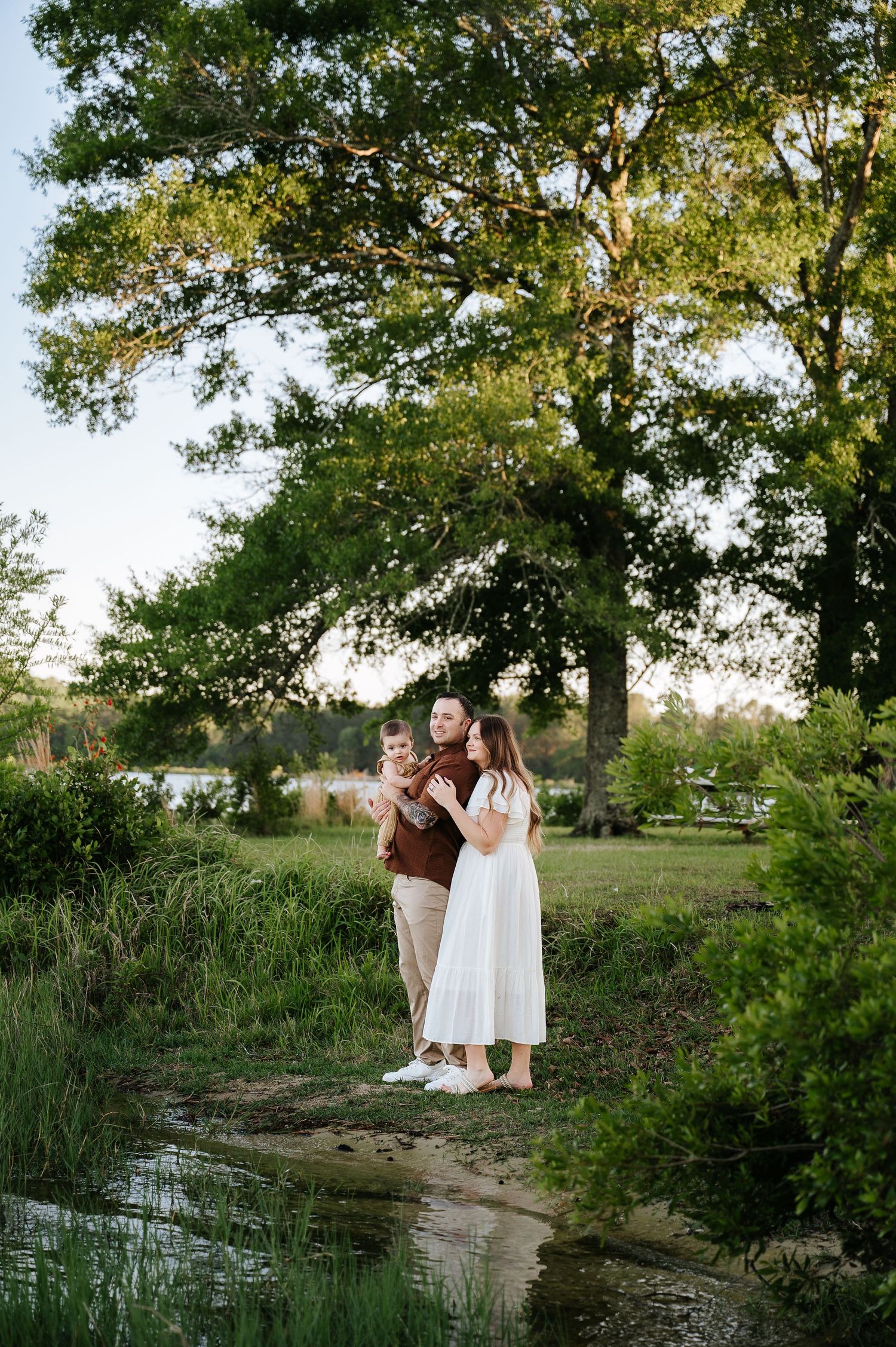 Family of three posing outdoors near a creek surrounded by lush green trees during golden hour.