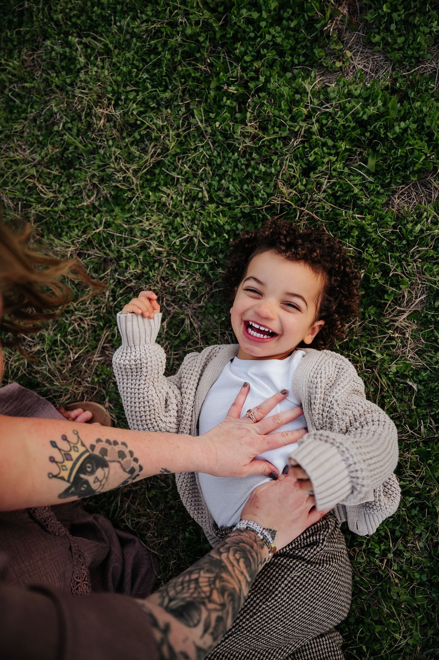 Hands with tattoos tickle a laughing child wearing a cozy knit sweater while lying on grass.