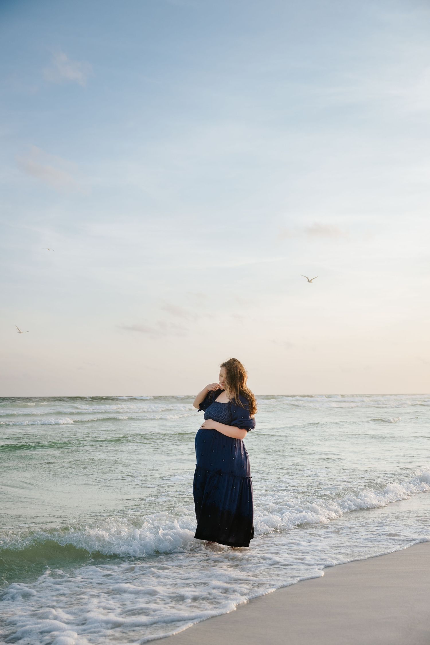 Pregnant woman in navy blue dress standing in ocean waves at sunset on a serene beach.