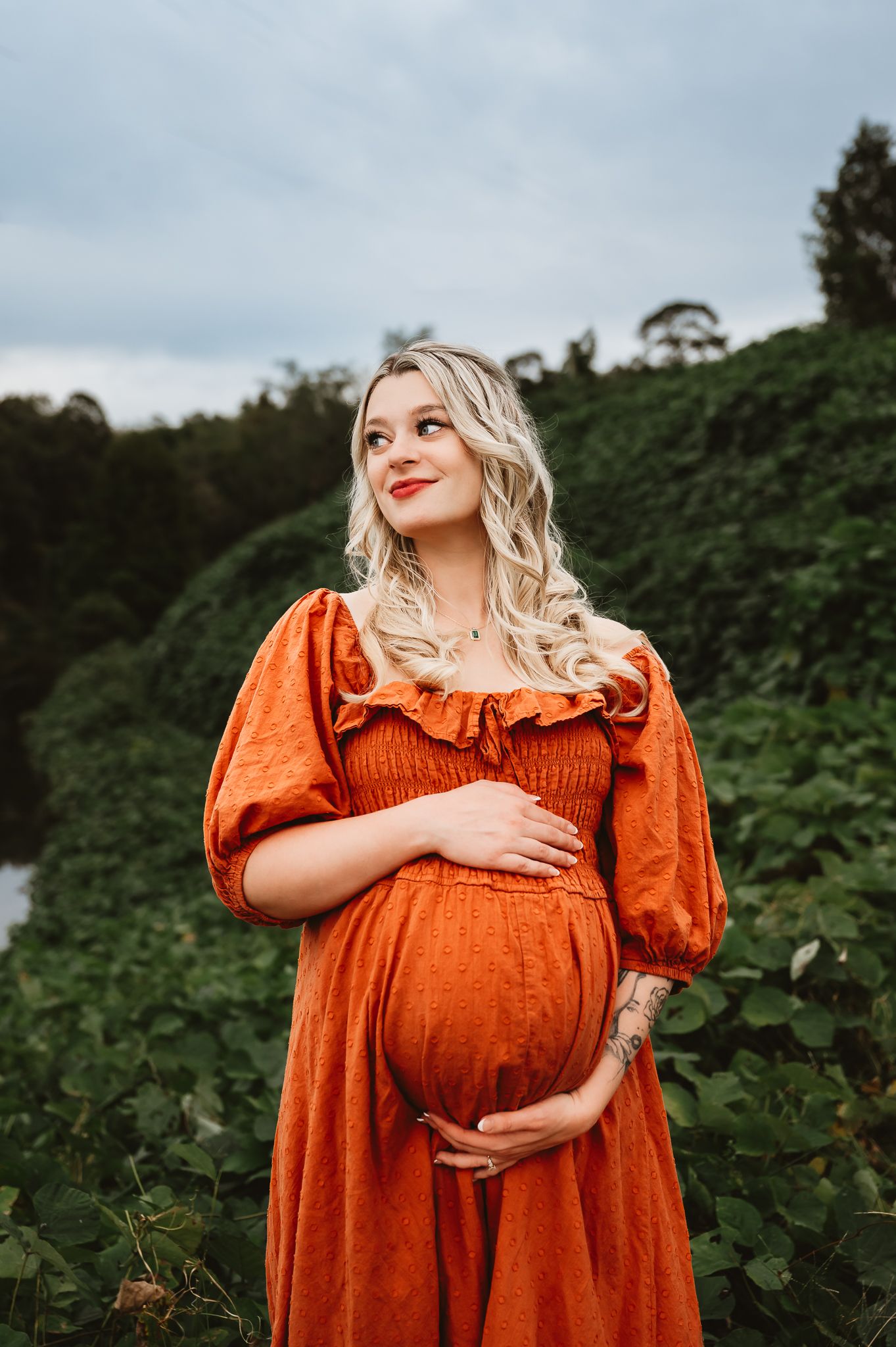 Pregnant woman in orange dress cradling baby bump outdoors among lush greenery.