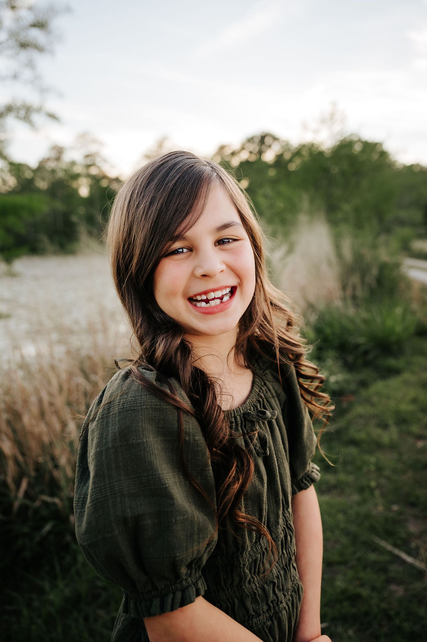 Smiling young girl with long brown hair wearing a green dress outdoors in a natural setting.