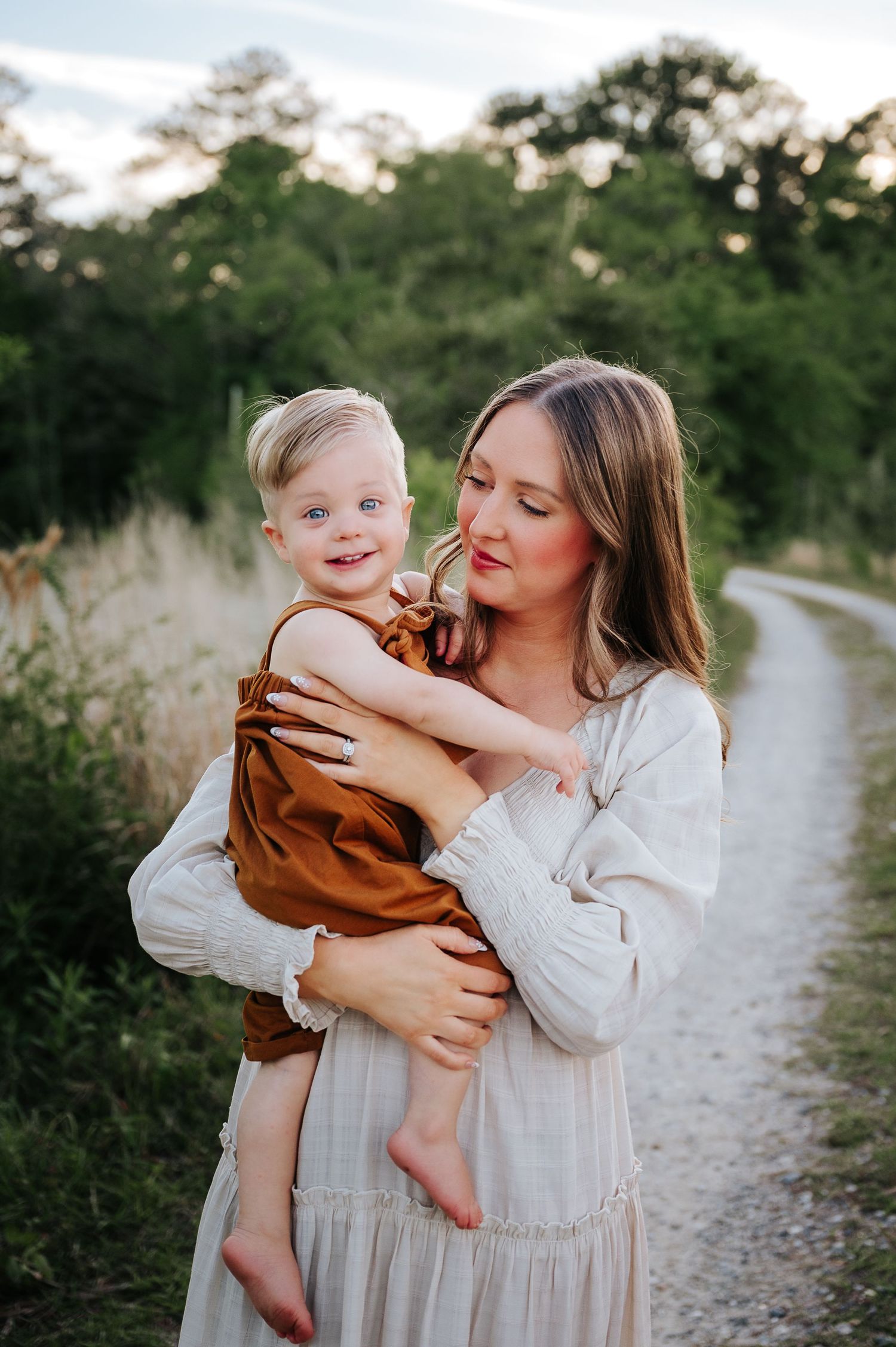 A mother in a white dress tenderly holds her smiling toddler on a scenic dirt path surrounded by lush greenery.