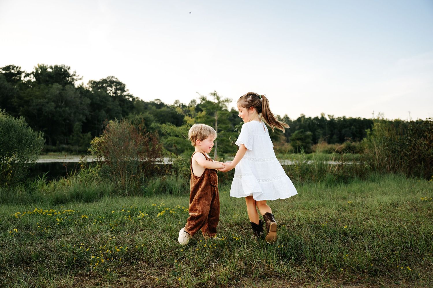 Two young children holding hands and playing in a lush green meadow near a pond at golden hour.
