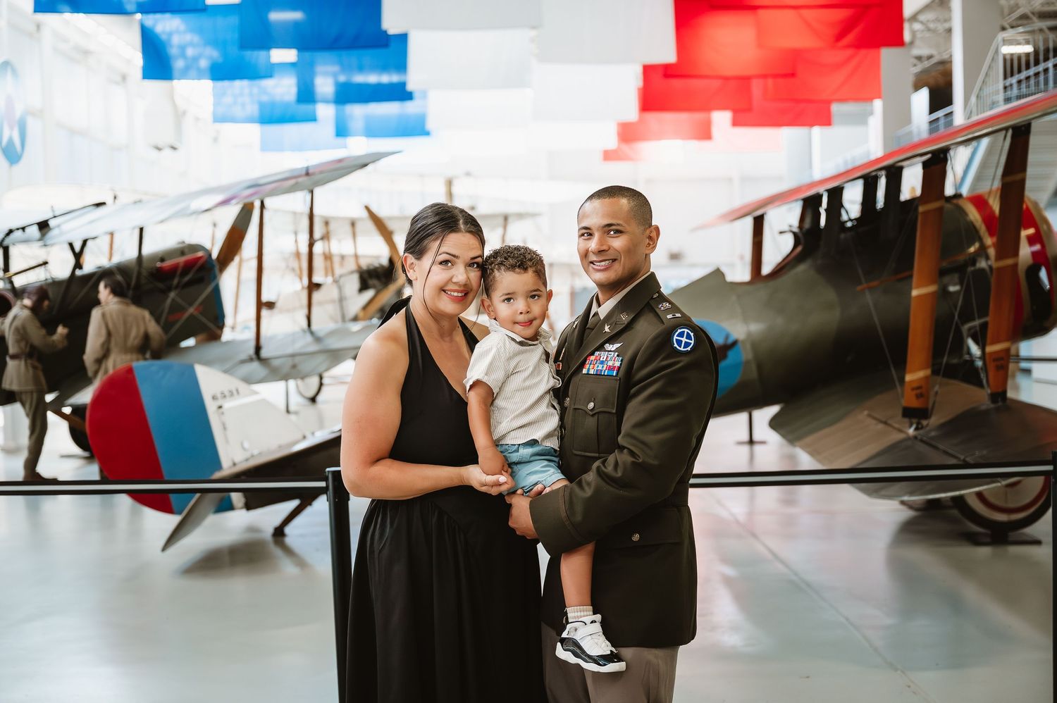 Military family poses with vintage biplane in aviation museum, man in uniform holds toddler beside woman in black dress.