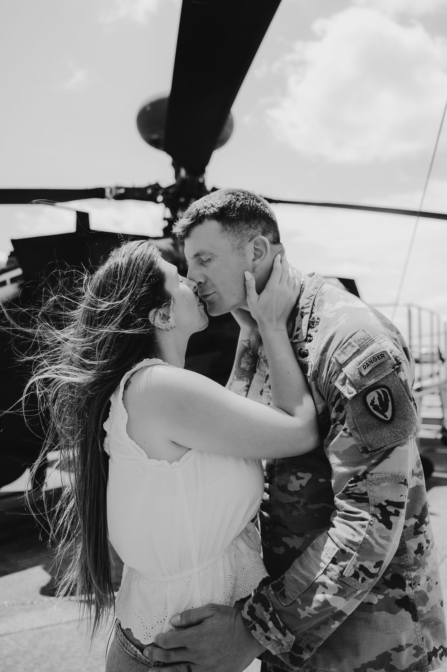 Black and white photo of a soldier in camouflage uniform sharing a romantic kiss with a woman near a military helicopter.