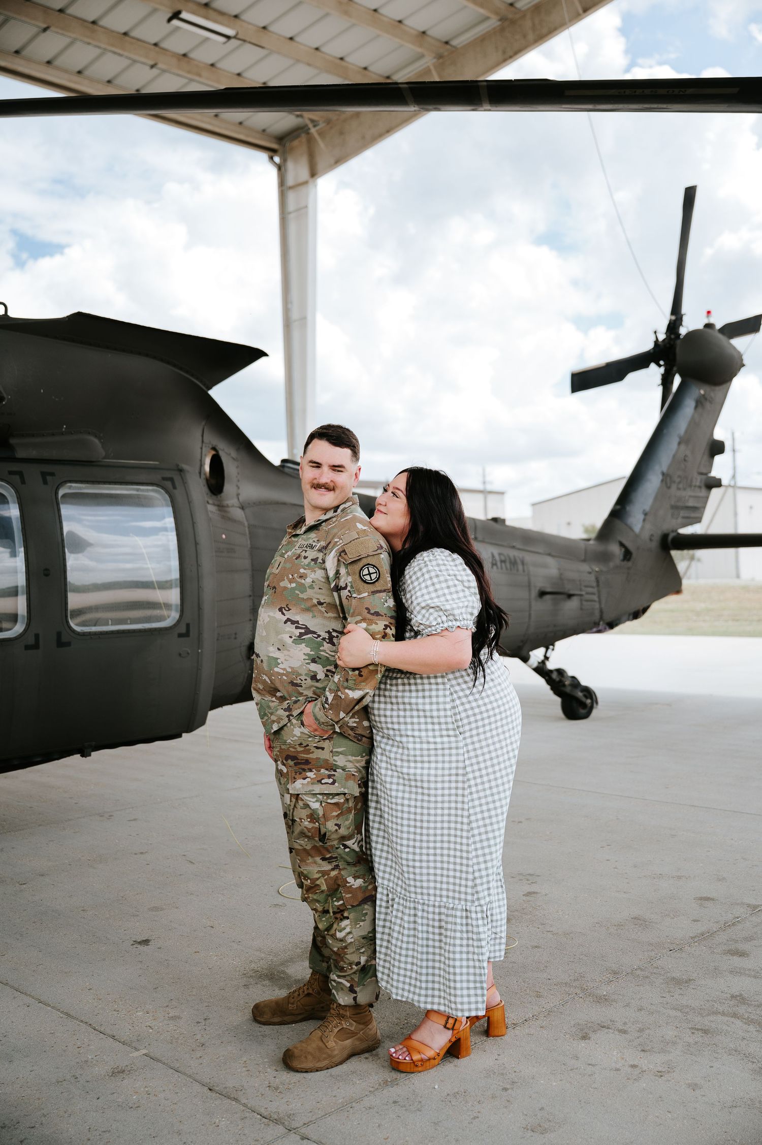 Military couple embracing in front of a Black Hawk helicopter in a hangar, soldier in camo uniform.