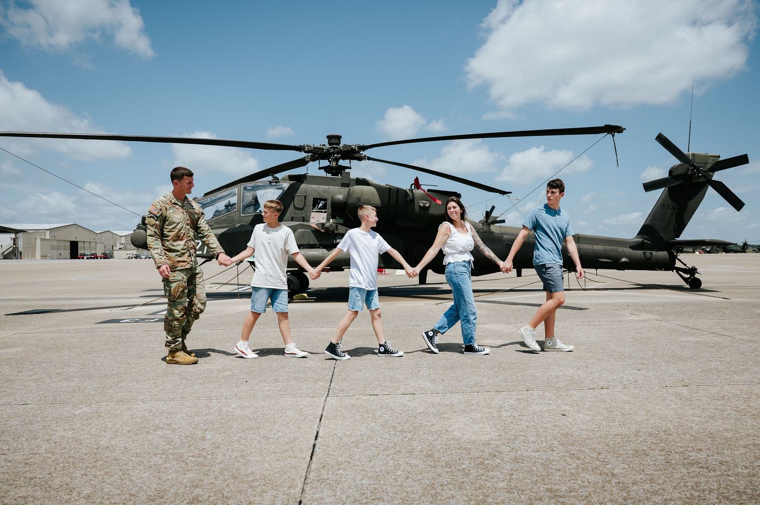 Military soldier holds hands with family in front of Apache helicopter on sunny airfield.