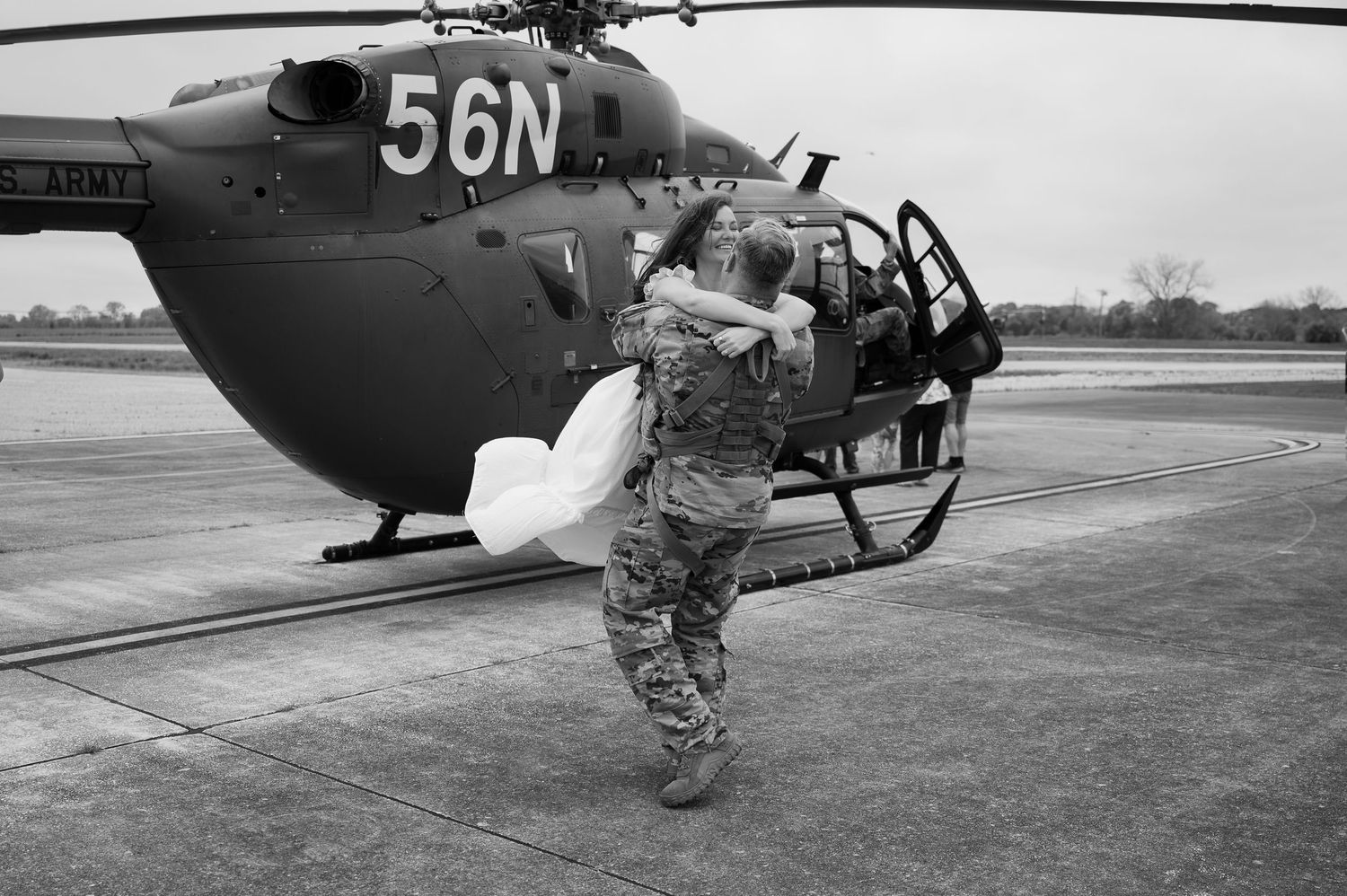 Soldier in camouflage embraces someone near military helicopter 56N on tarmac in black and white photo.