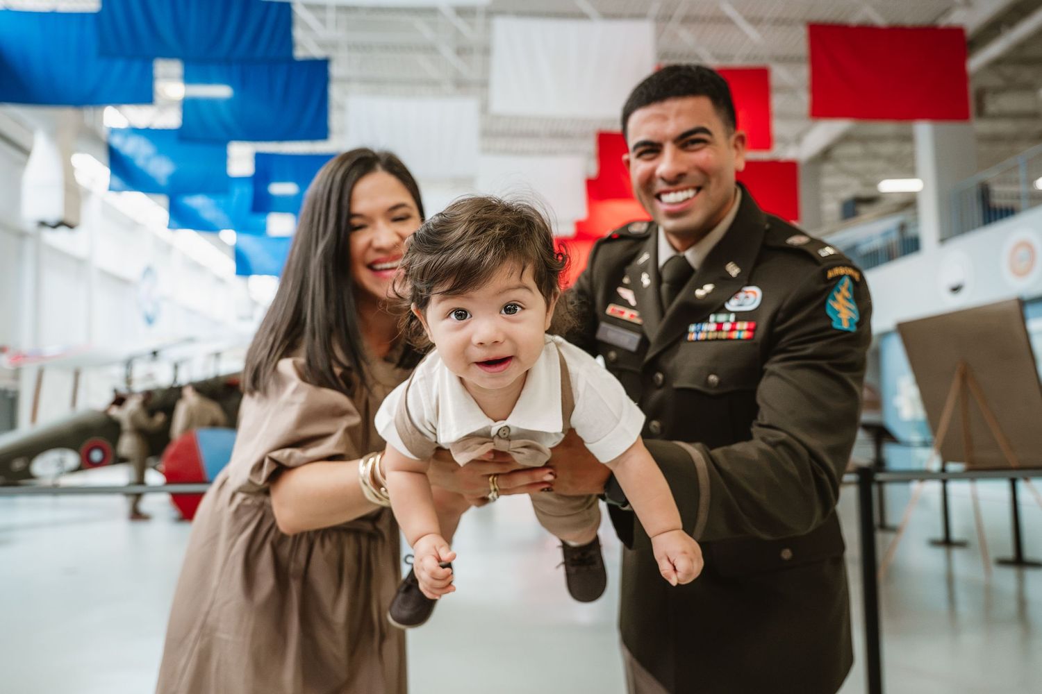 Happy military family with soldier in dress uniform holding smiling baby at aviation museum.