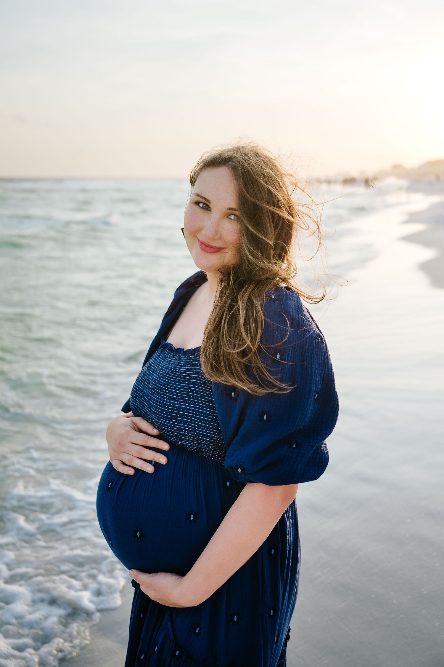 Pregnant woman in blue dress cradling baby bump on beach at sunset with ocean waves behind her.