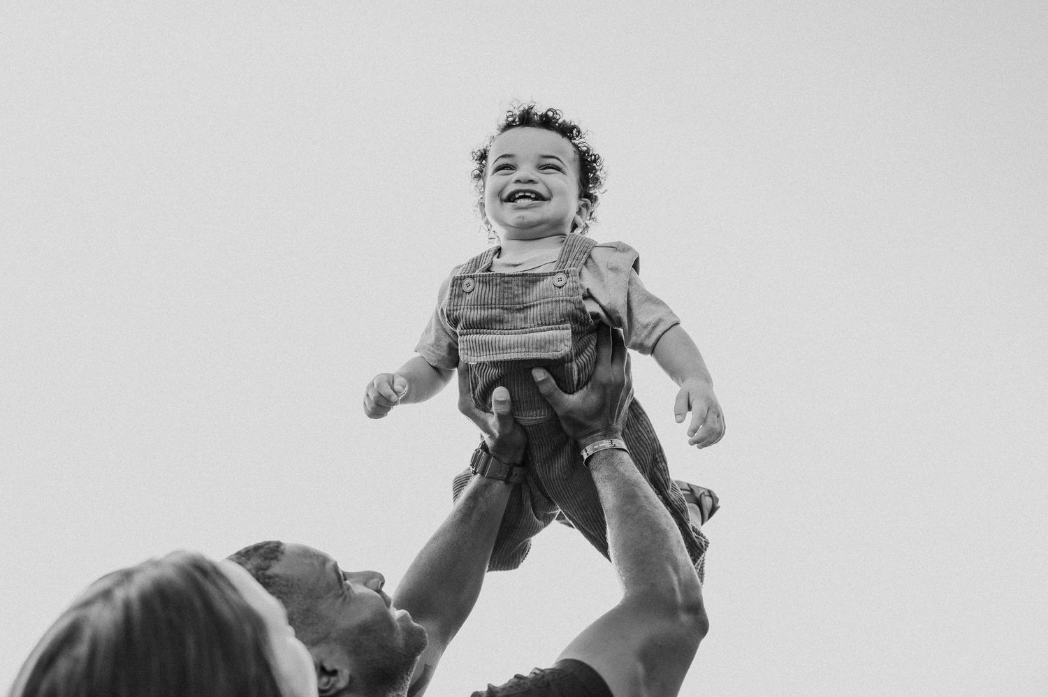 A black and white photo capturing pure joy as a toddler is lifted high in the air outdoors.