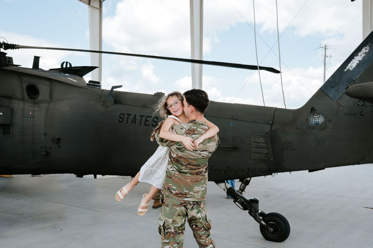 Soldier in camouflage uniform joyfully lifting partner in white dress during military homecoming near helicopter.