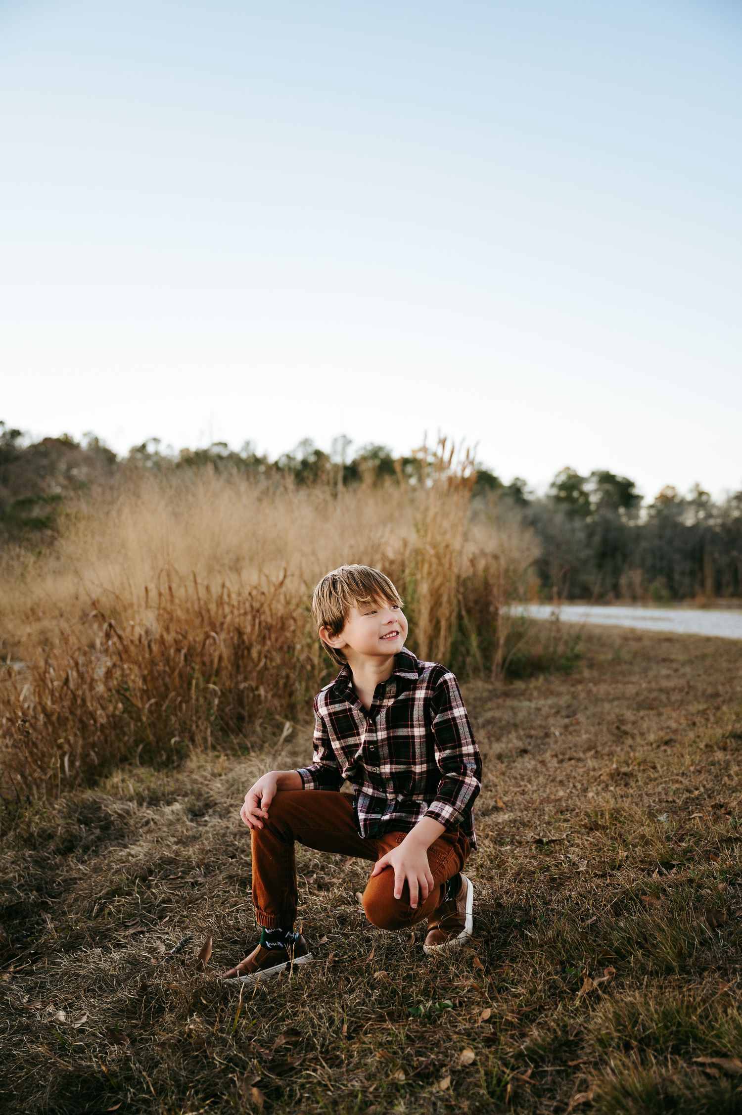 A person in a plaid flannel shirt crouches in a field of tall grass during golden hour.