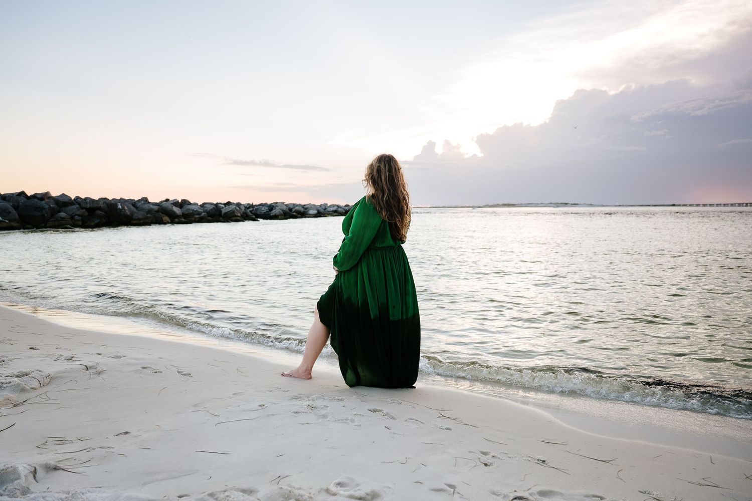 Woman in green dress sitting on white sand beach at sunset with calm ocean waters and rocky jetty in background.