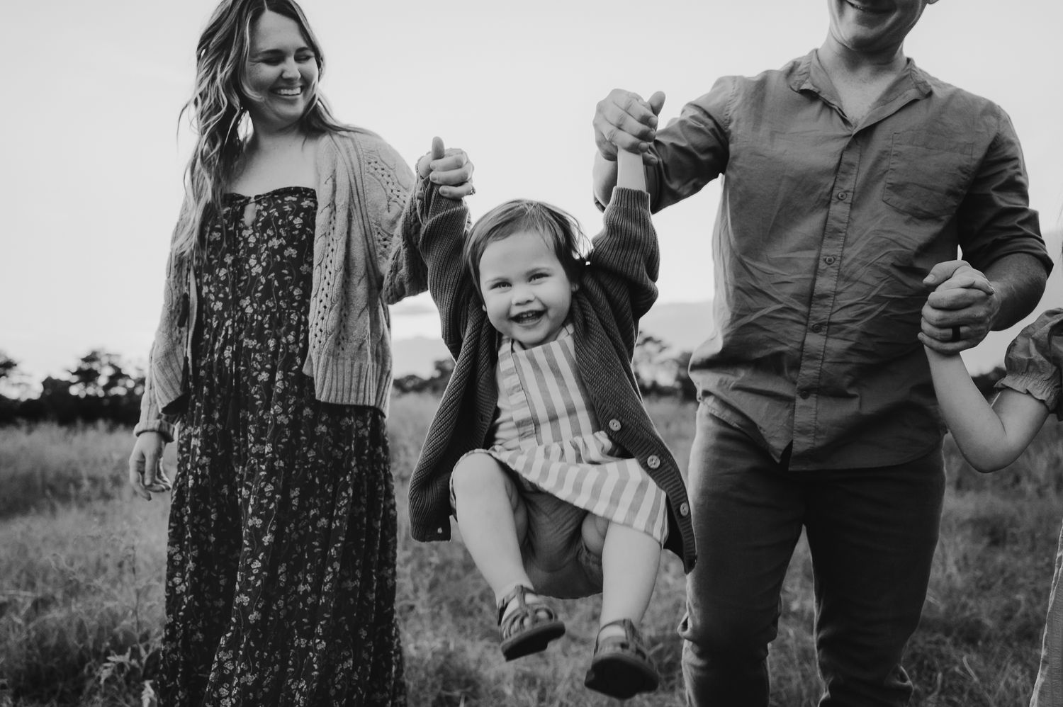 A family in a grassy field swings their young child between them while walking in black and white photography.