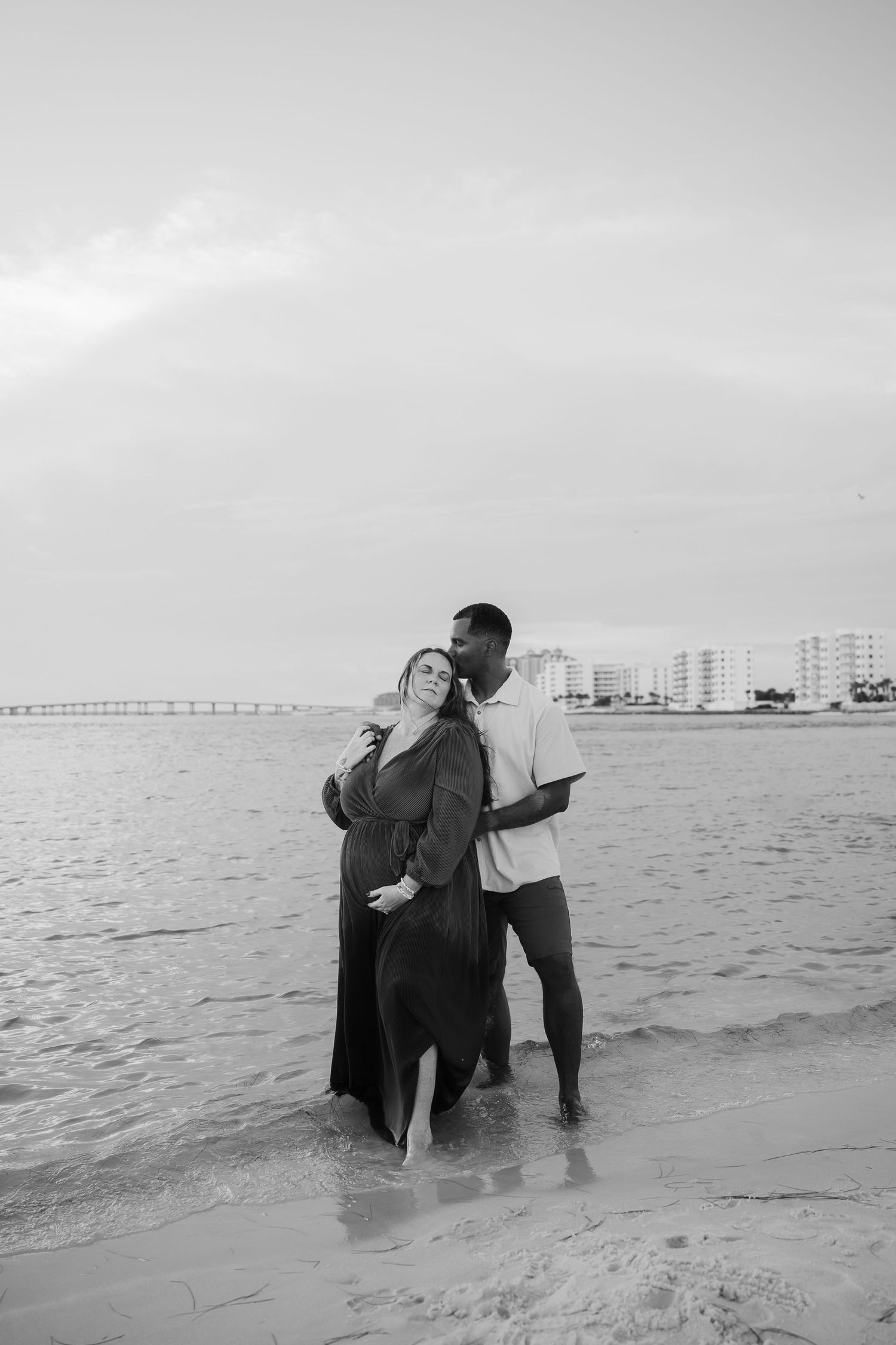 Black and white beach maternity photo of a couple standing in ocean waves with city skyline in background.