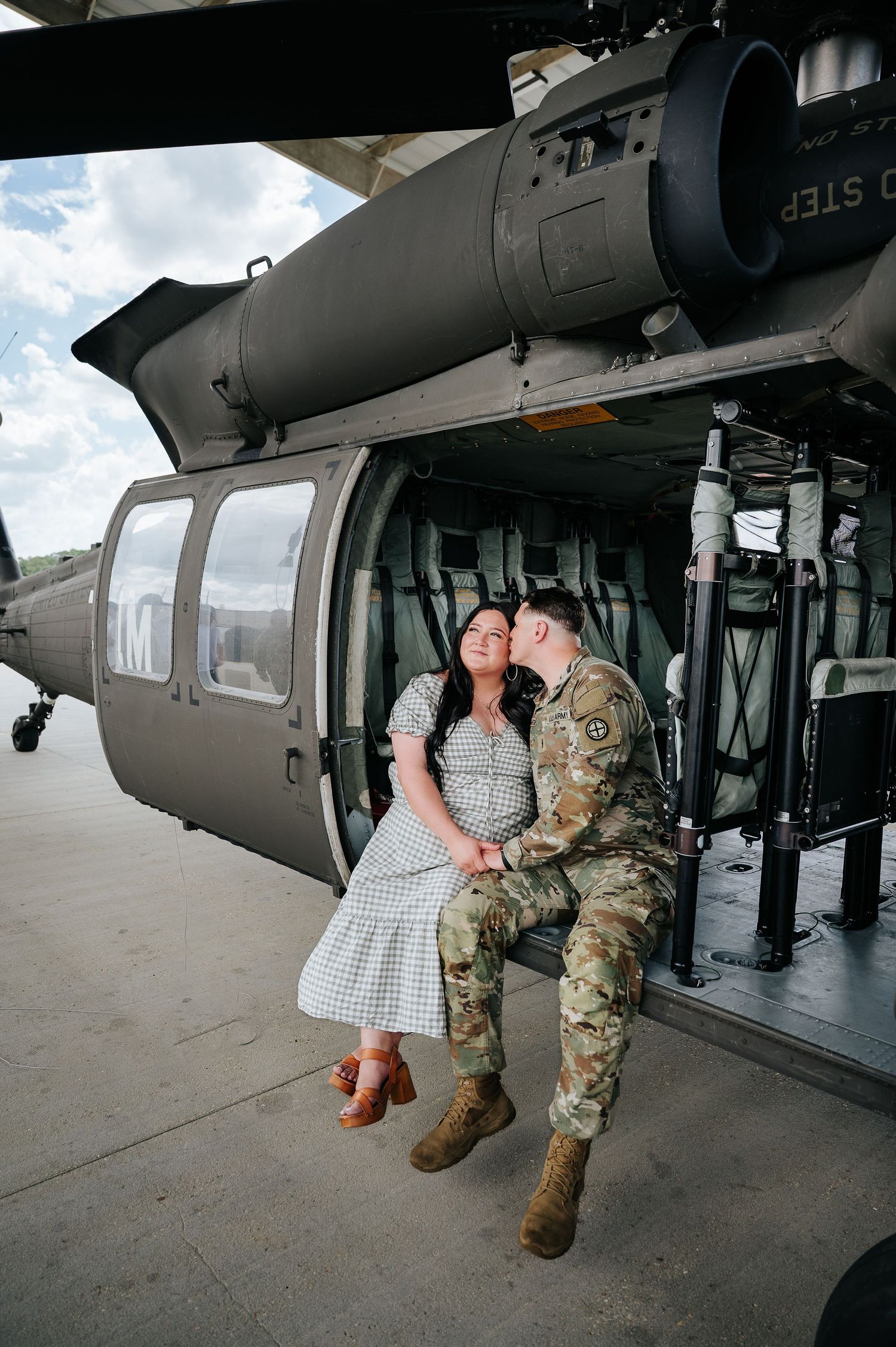 A soldier in camouflage uniform kisses a woman in a gingham dress beside a military helicopter on a tarmac.