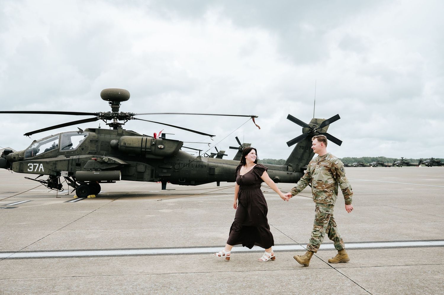 A couple holds hands on a military airfield with an Apache helicopter in the background.